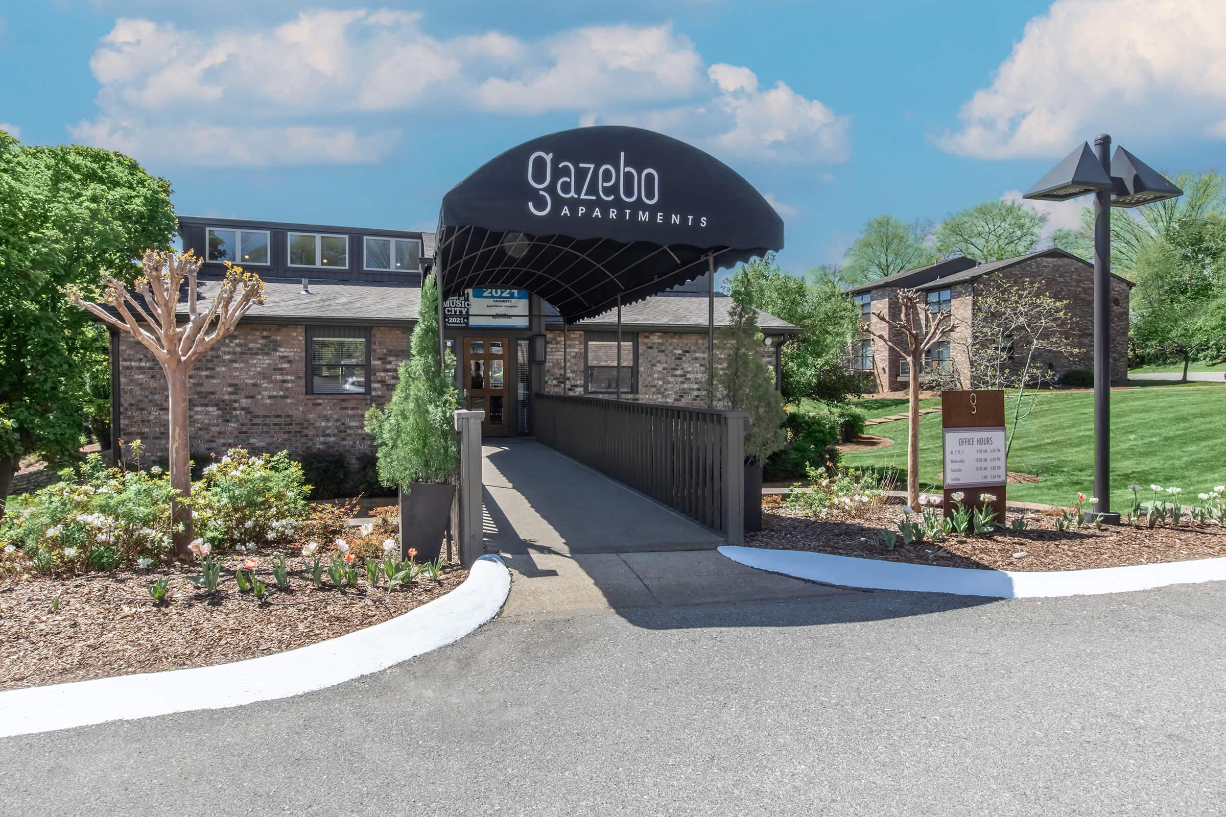 Entrance to Gazebo Apartments featuring a welcoming awning and landscaped surroundings. The pathway leads to the building, flanked by greenery and flowerbeds. Signs indicate directions and information about the apartments, with a clear blue sky in the background.