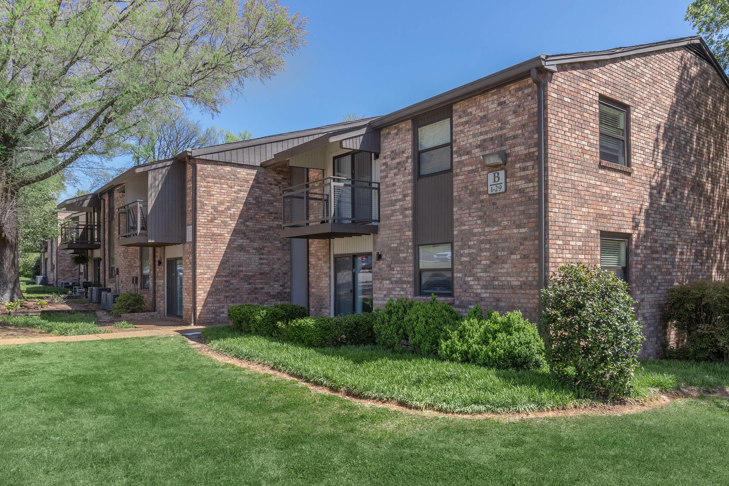 A modern brick apartment building with multiple units, featuring balconies and large windows. The landscaping includes a well-kept lawn, bushes, and trees, with a clear blue sky in the background.