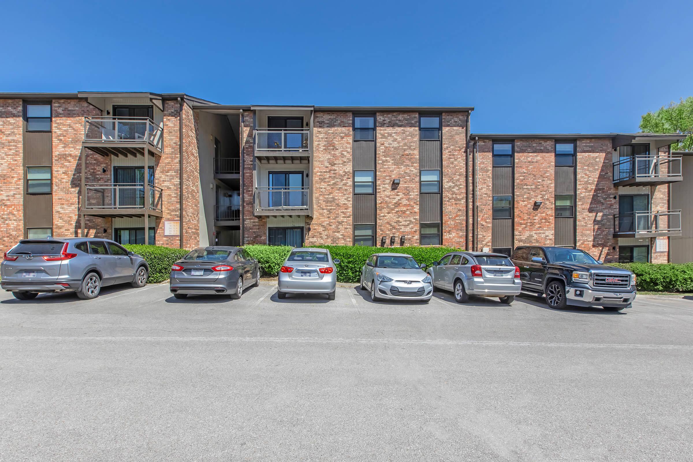 A modern apartment complex with a brick facade and multiple balconies. Several parked cars of various makes and models are lined up in front of the building on a clear, sunny day with a blue sky. Lush greenery borders the parking area.