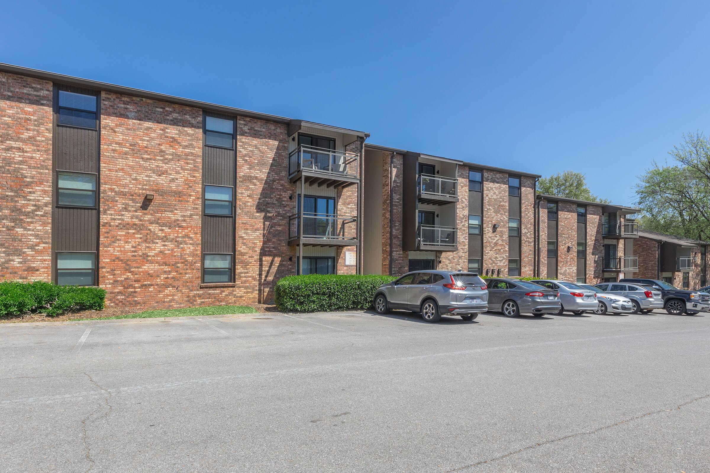 A multi-story brick apartment building with black accents, featuring balconies on the second floor. The foreground shows a parking lot with several cars parked along the side of the building, and trees in the background under a clear blue sky.
