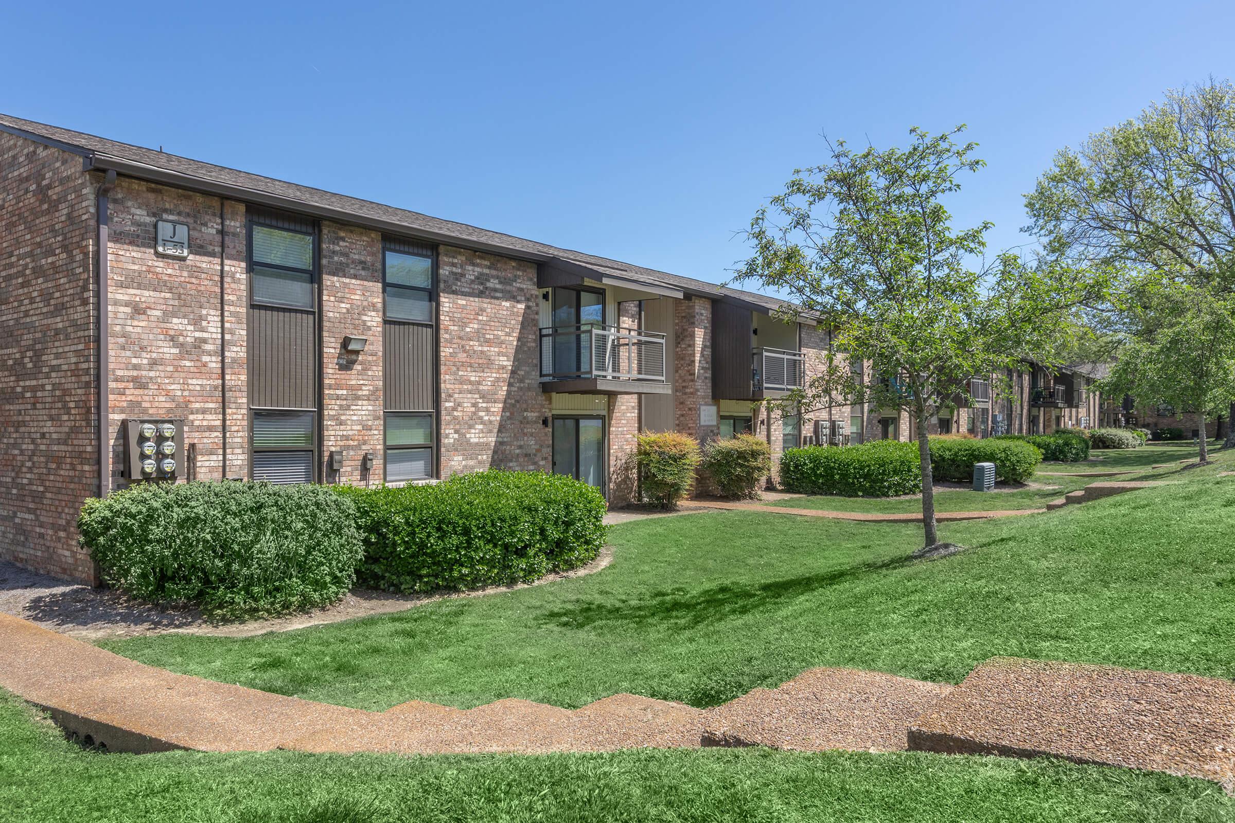 A view of a multi-unit residential building with brick exteriors. Lush green lawns and landscaped shrubs line the walkways. The sky is clear blue, and there are steps leading up to the entrance of the units. The setting appears inviting and well-maintained, typical of an apartment complex.