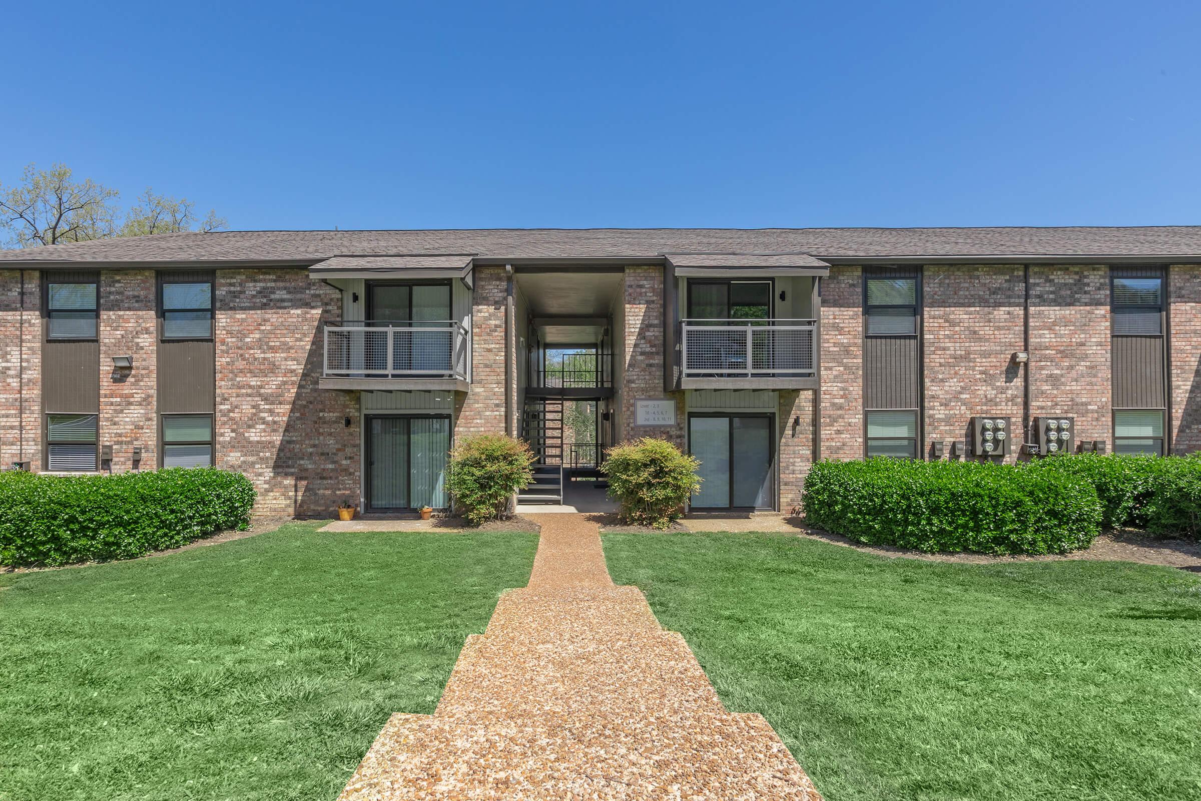 A modern two-story brick apartment building featuring a well-maintained lawn, a gravel pathway leading to the entrance, large windows with balconies, and air conditioning units mounted on the exterior walls. The sky is clear and blue, adding to the vibrant atmosphere of the scene.