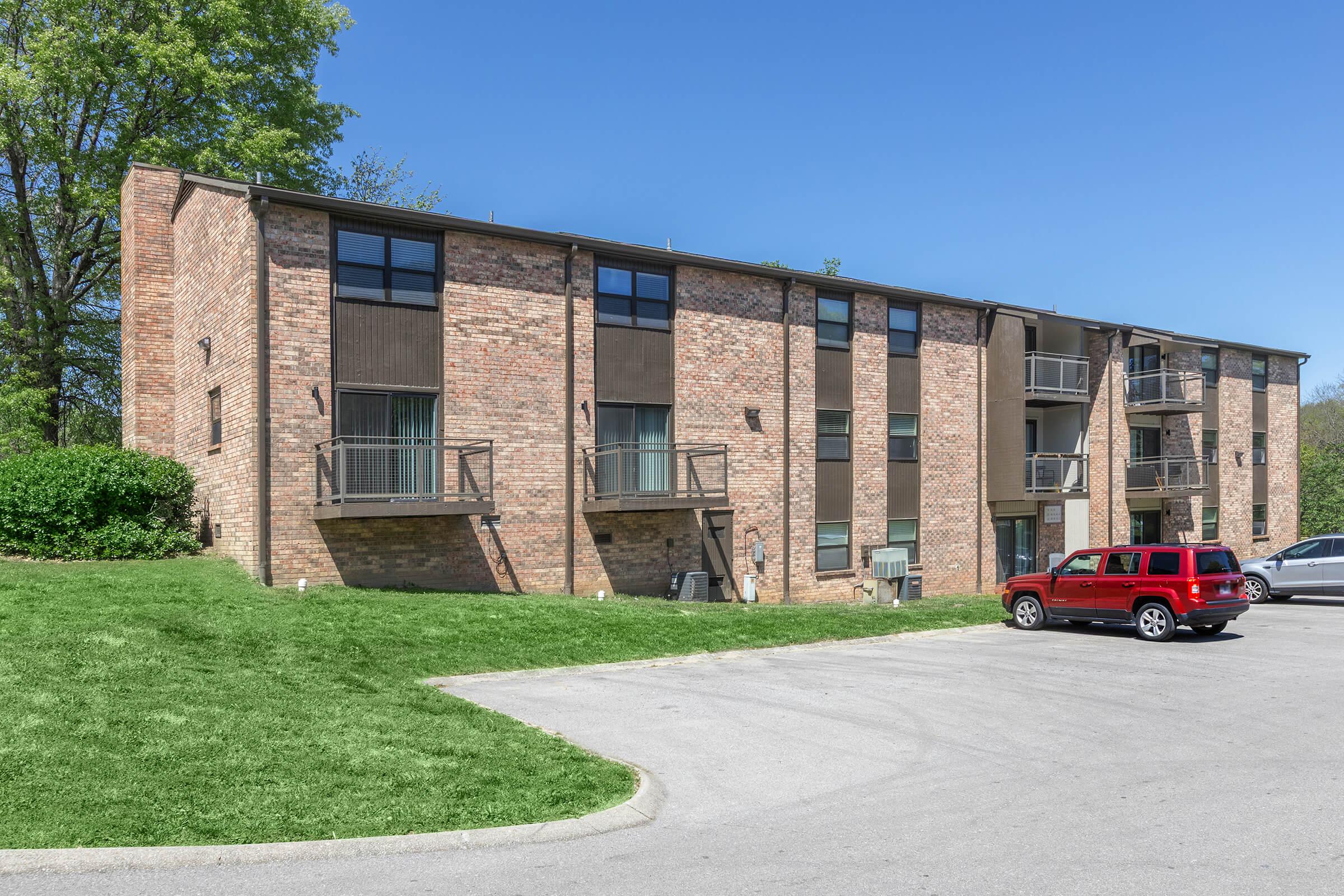 Two-story brick apartment building with multiple balconies and a grassy area in front. A red SUV is parked in the driveway, and several trees are visible in the background under a clear blue sky.