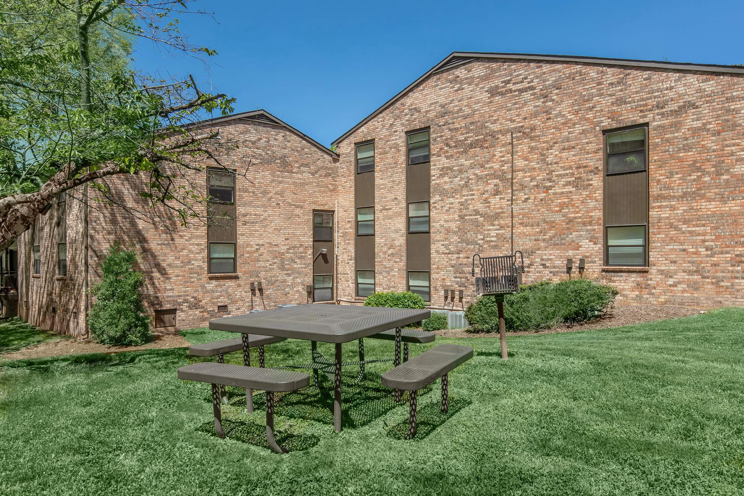 A well-maintained outdoor area featuring a gray picnic table with benches set on a green lawn. In the background, there are two brick buildings with large windows and a small tree. The sky is clear and blue, creating a pleasant atmosphere for relaxation or gatherings.