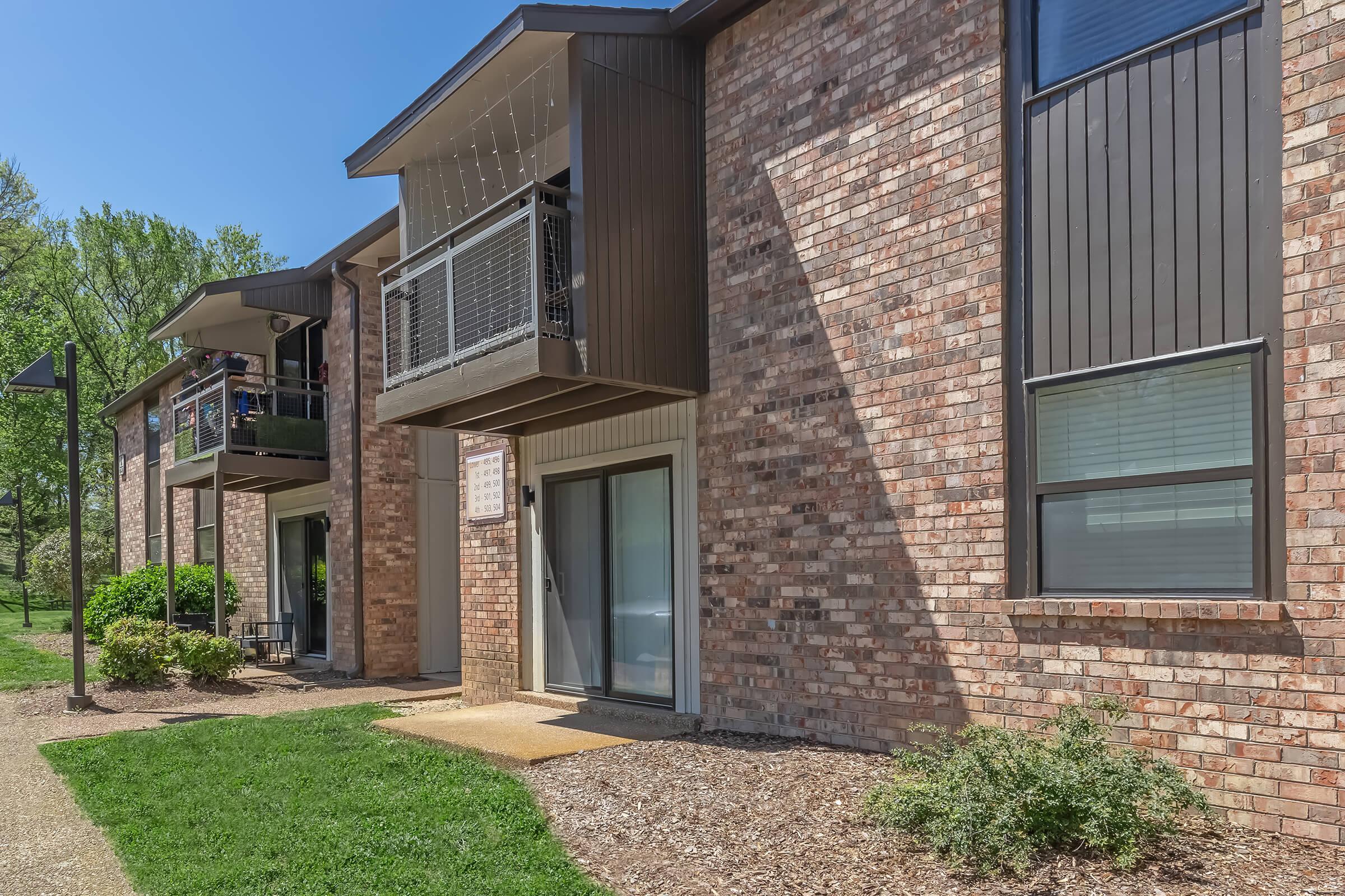 A modern brick apartment building with two stories, featuring balconies and large windows. The foreground includes well-manicured grass and landscaping, with a clear blue sky above. The building has a contemporary design, showcasing a mix of brick and modern architectural elements.