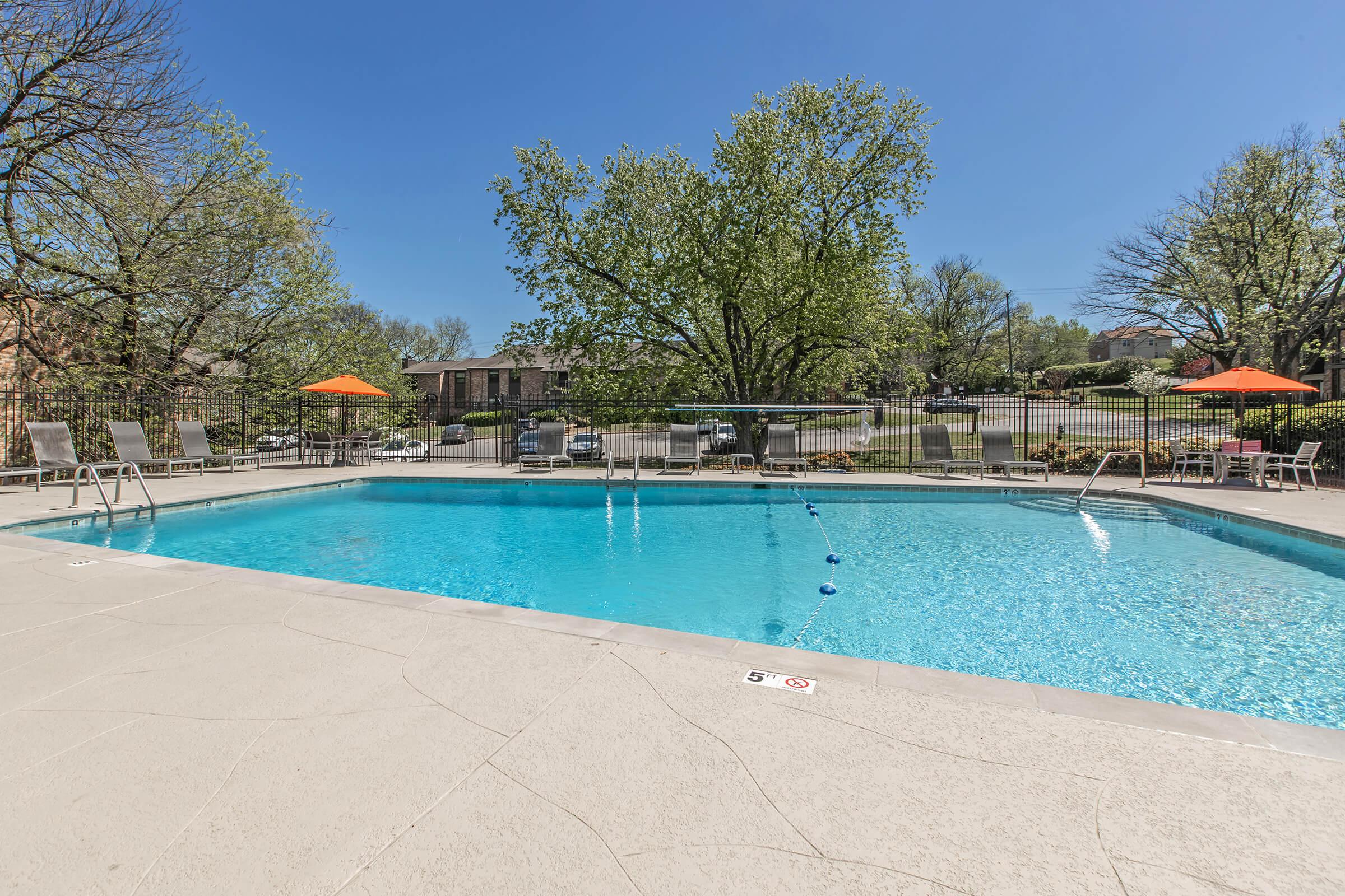 A clear blue swimming pool surrounded by lounge chairs and orange umbrellas. Tall trees provide shade in the background, and a bright blue sky enhances the inviting outdoor atmosphere. The pool area is well-maintained, suggesting a relaxing spot for leisure and recreation.