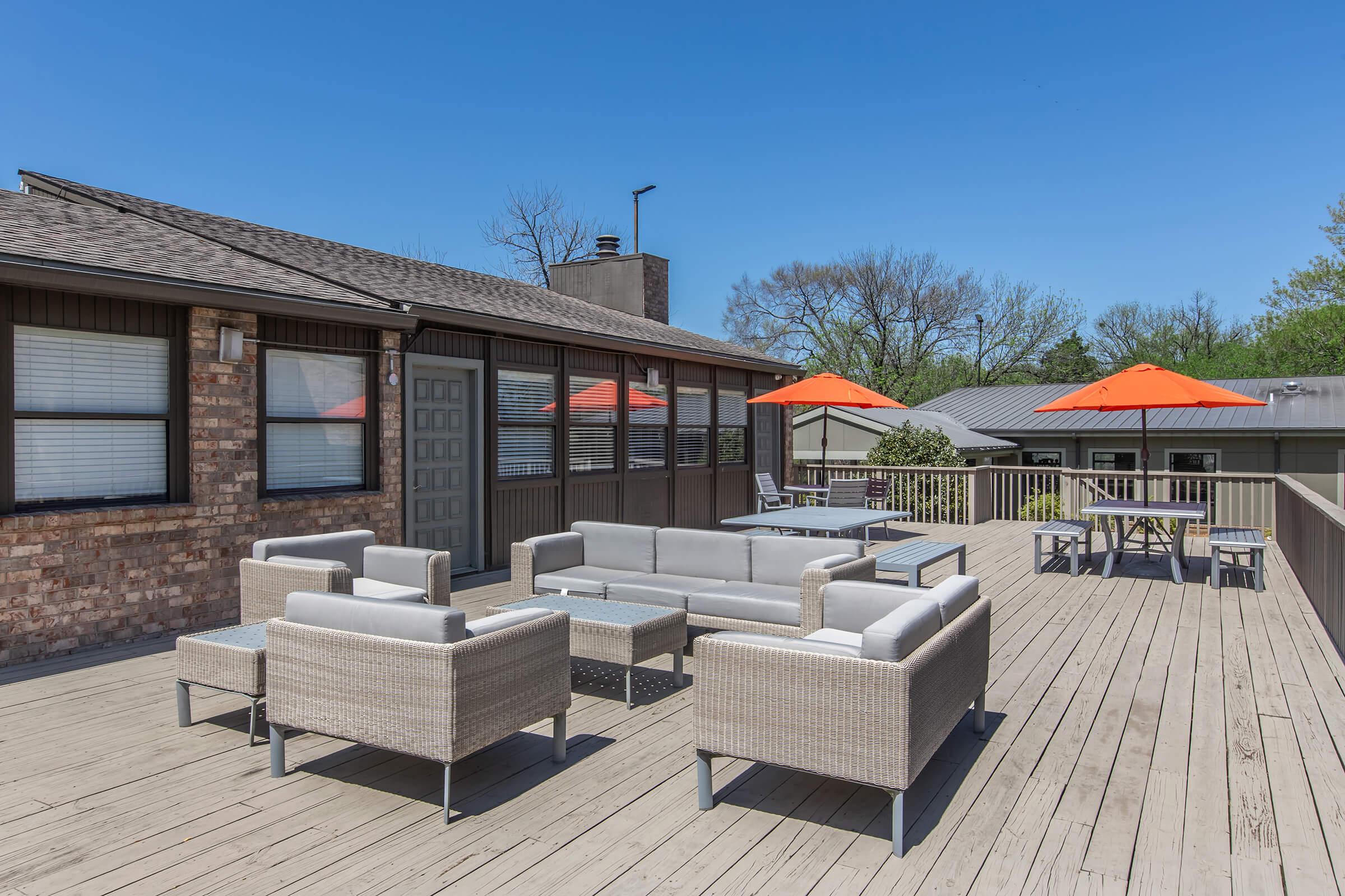 A spacious rooftop patio with modern gray wicker seating arranged around a coffee table. Bright orange umbrellas provide shade over tables and chairs. The area features a wooden decking, surrounded by greenery and blue skies, creating a pleasant outdoor space for relaxation or social gatherings.
