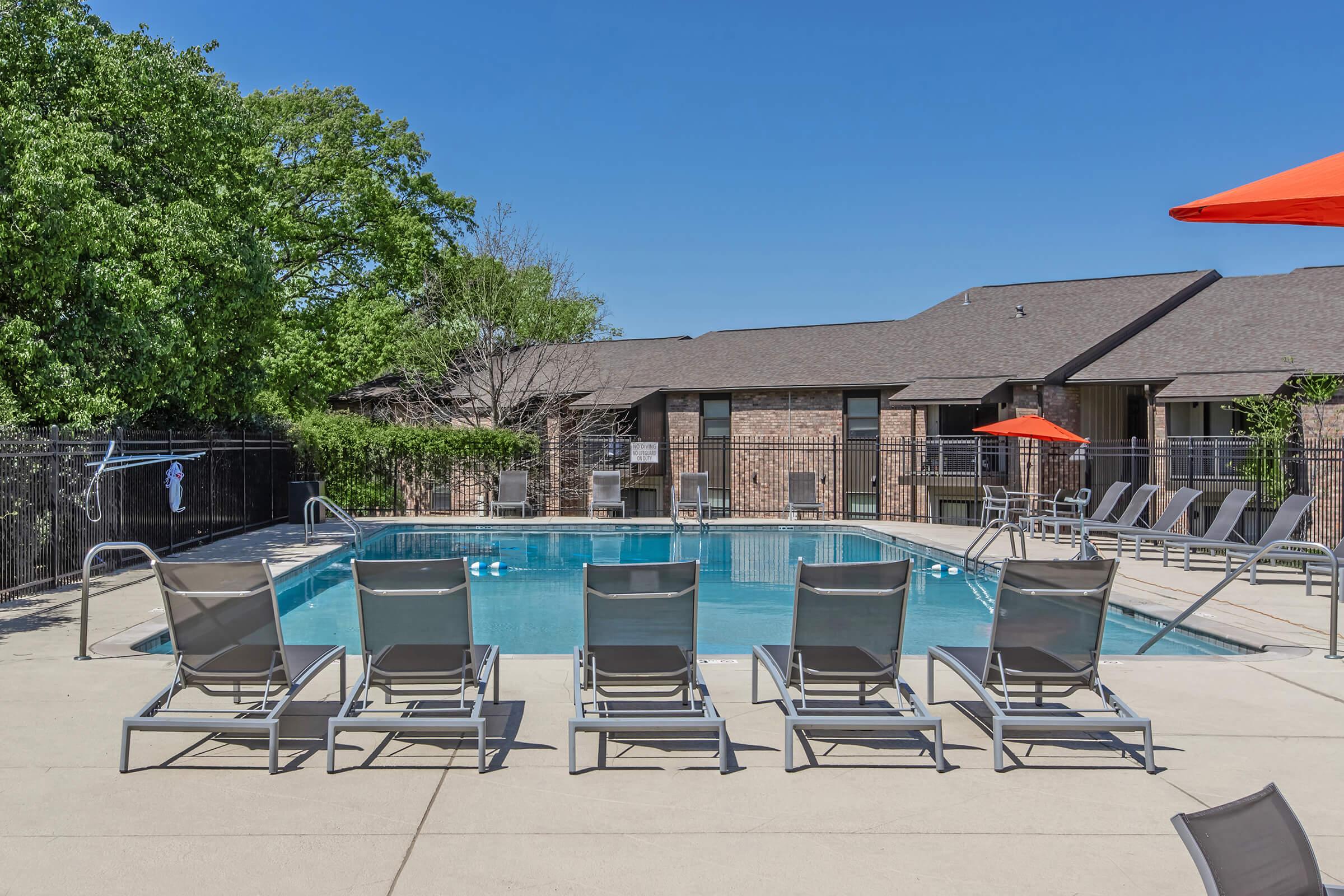A swimming pool with lounge chairs arranged around it, set in a sunny outdoor area. The pool reflects the clear blue sky, and there's an orange umbrella providing shade. Surrounding the pool are green trees, a fence, and a brick building in the background, creating a relaxing atmosphere.