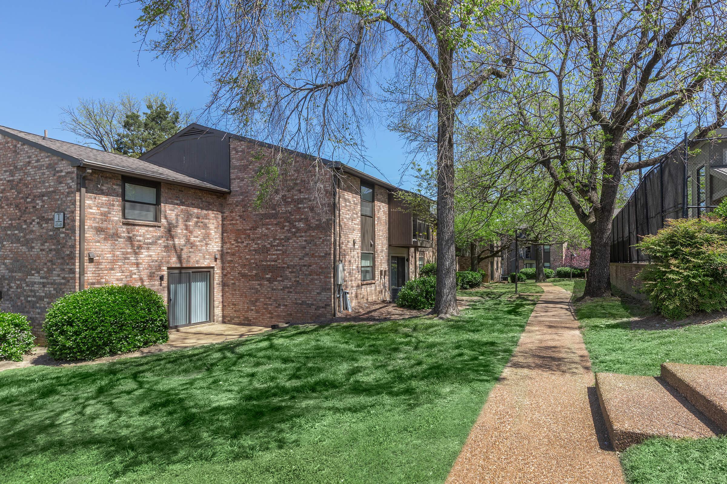 A scenic view of a residential area featuring two brick apartment buildings surrounded by well-maintained green grass and trees. A pathway lined with gravel leads through the area, providing access to the buildings and outdoor spaces on a clear, sunny day.