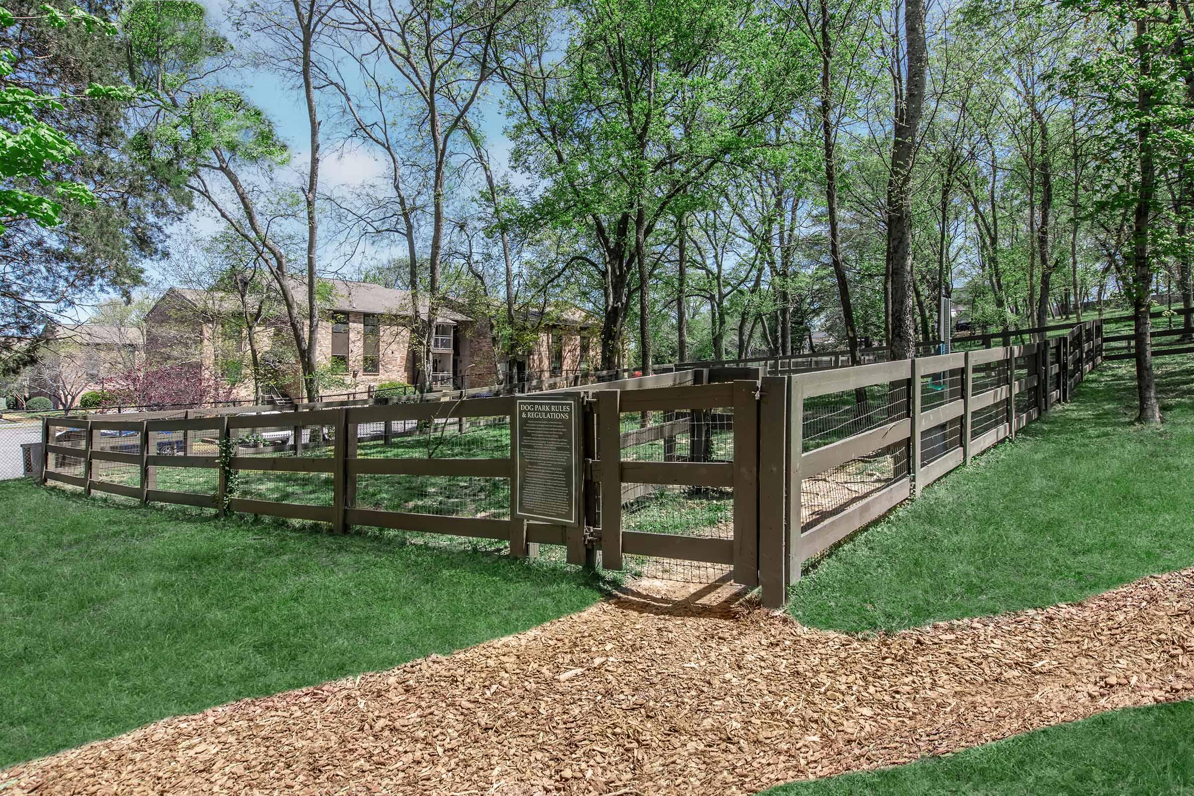 A fenced area surrounded by trees and grass, with a path made of wood chips leading towards a sign. In the background, a building is partially visible through the trees, indicating a residential or community space. The scene is bright and well-maintained, reflecting a pleasant outdoor environment.