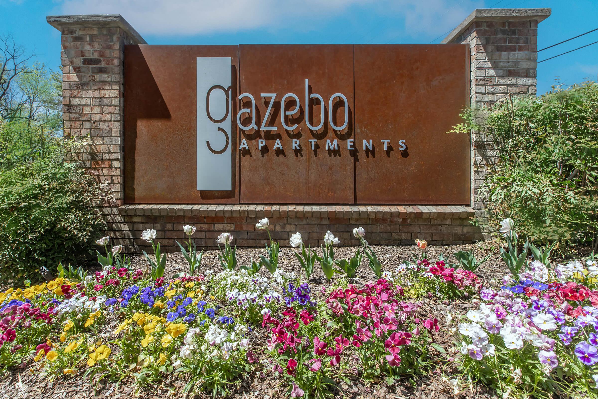 Sign for Gazebo Apartments featuring modern lettering. Surrounding the sign is a colorful flower bed with various blooms, including tulips and pansies. The background shows a clear blue sky, adding to the inviting atmosphere of the apartment complex.