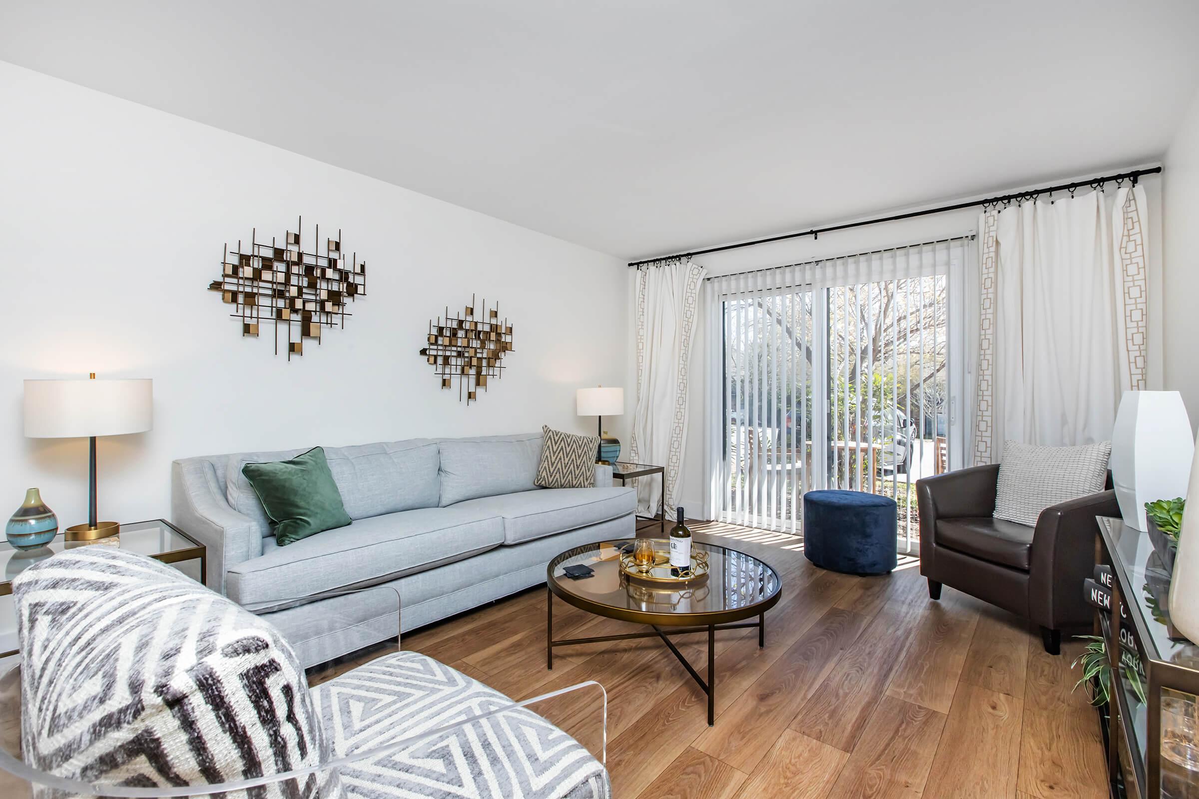 A modern living room featuring a light gray sofa with decorative pillows, a round coffee table, and two unique accent chairs. Natural light streams in through sliding glass doors with sheer curtains, revealing a view of outdoor greenery. Wall art adds a contemporary touch to the decor.
