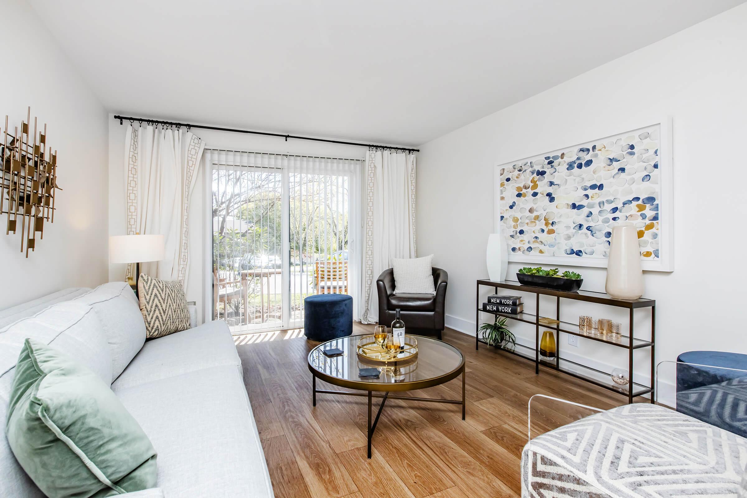 A modern living room featuring a light blue sofa with decorative pillows, a round glass coffee table, and stylish accessories. There's a cozy armchair, a large wall art piece with blue and yellow patterns, and large windows that let in natural light. The wooden flooring adds warmth to the space.