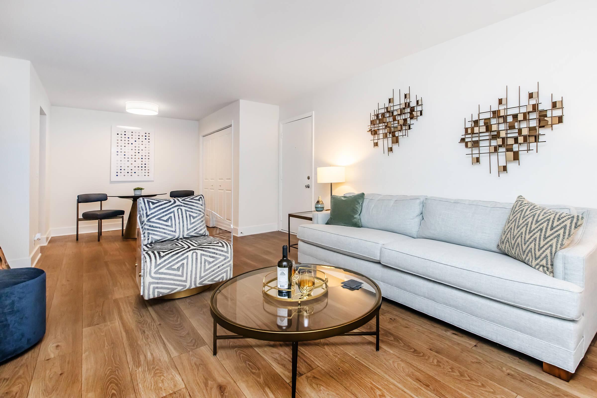 A bright and modern living room featuring a light gray sofa with decorative cushions, a round coffee table with a wine bottle and glass, a patterned accent chair, and a dining area in the background with four chairs. Wall art consists of geometric metal sculptures, enhancing the contemporary decor.
