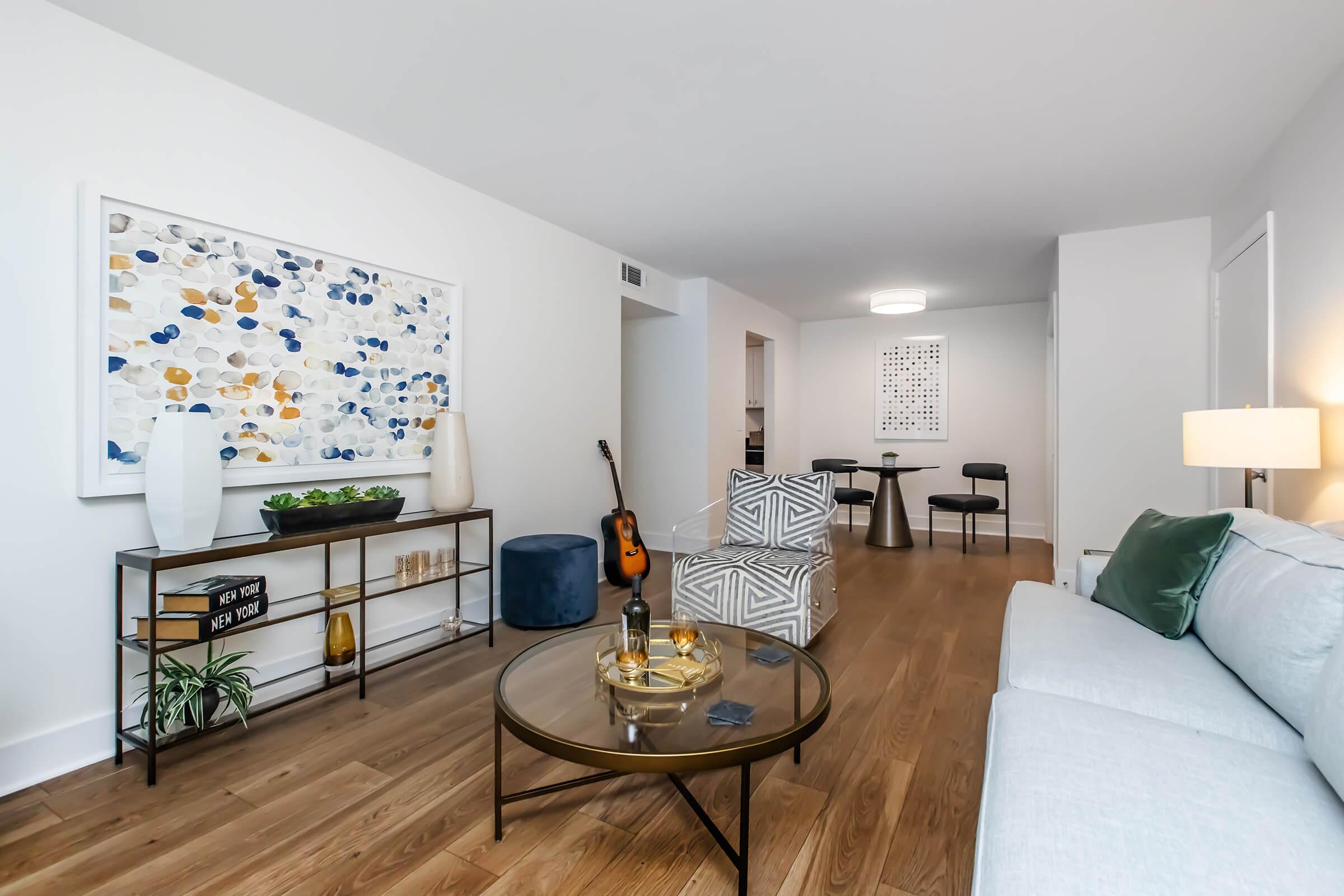 A modern living room featuring a light-colored sofa, a glass coffee table with decorative items, a guitar, and stylish decor. The walls are adorned with abstract art and plants, creating a contemporary and inviting atmosphere. A dining area and kitchen area are visible in the background.