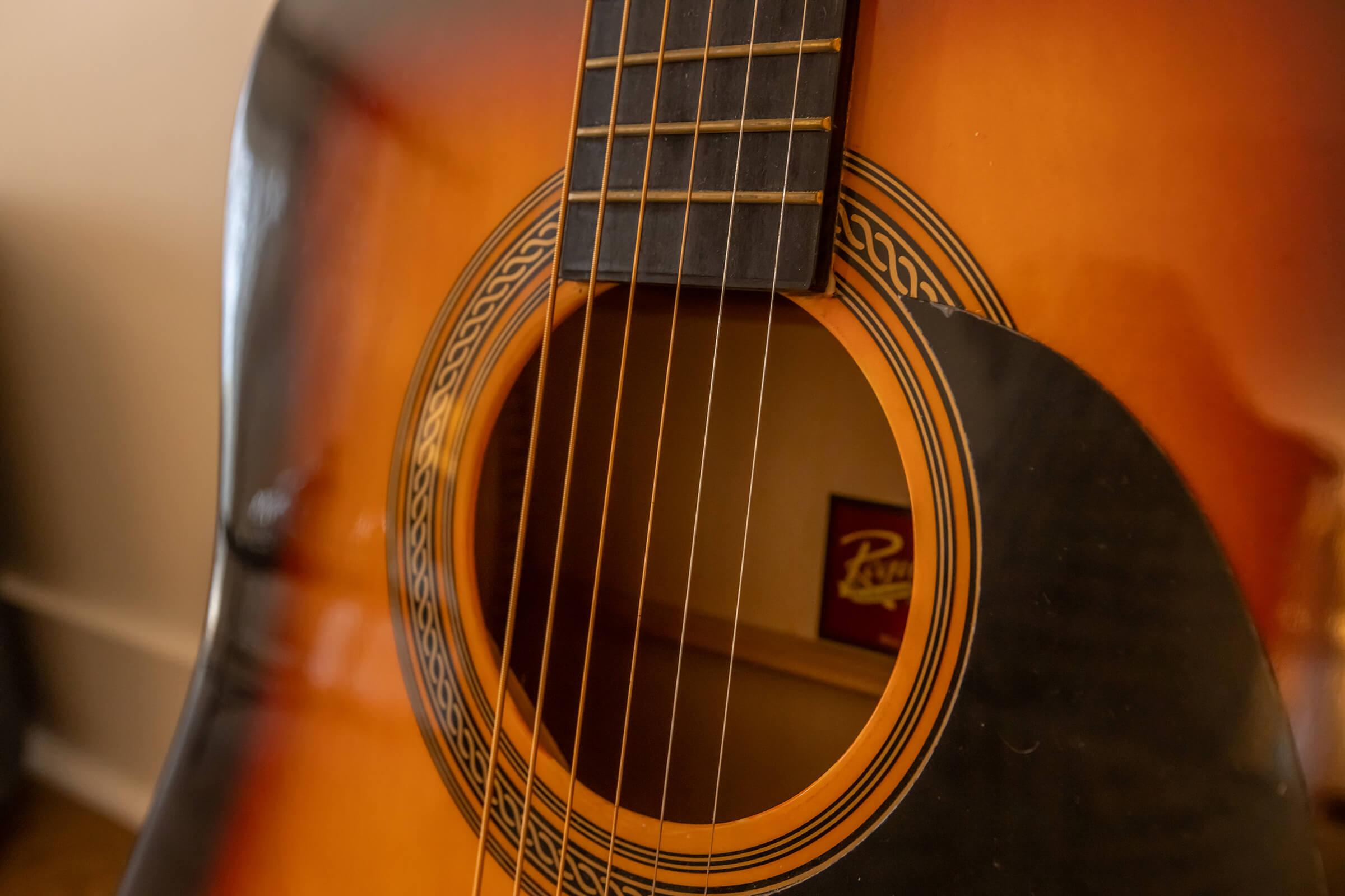 Close-up view of an acoustic guitar, focusing on the guitar's soundhole and neck. The wood grain and detailing around the soundhole are visible, along with the strings and fretboard. A blurred background suggests a cozy indoor setting.