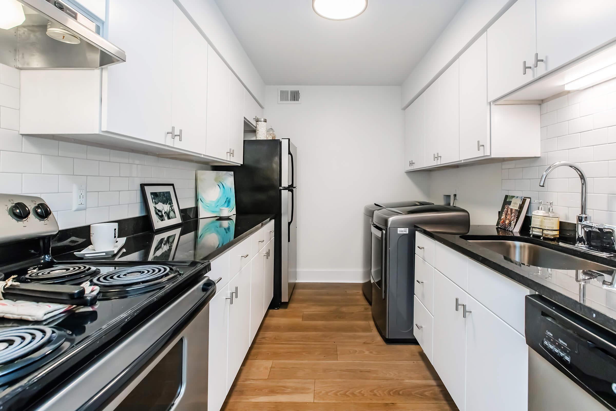 A modern kitchen featuring white cabinetry, stainless steel appliances, and black countertops. The space includes a stove, oven, refrigerator, and washing machine. Decorative items are displayed on the countertops, and the flooring is wooden. Bright lighting enhances the clean and airy atmosphere.