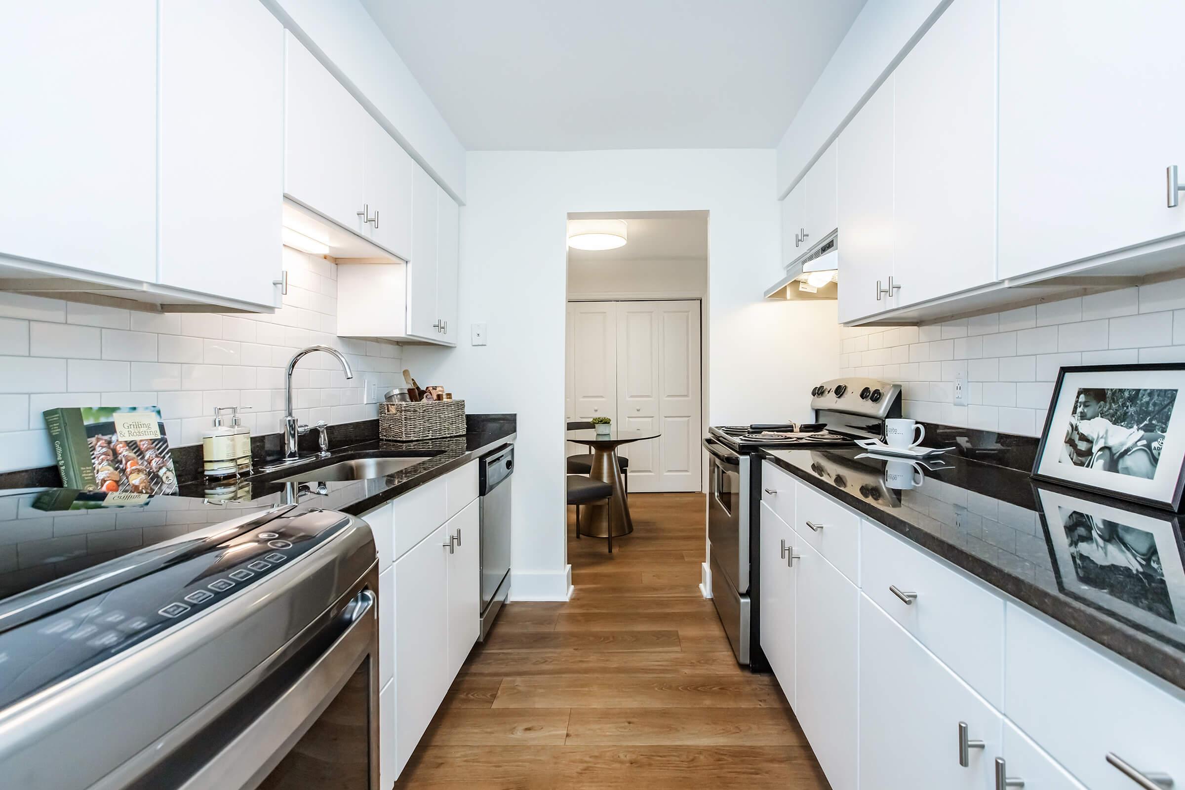 A modern kitchen featuring stainless steel appliances, including a washer and oven, sleek white cabinets, and a dark granite countertop. A hallway leads to another room, and there are decorative items like a framed photo and cookbooks on the counter, with warm wooden flooring completing the inviting look.