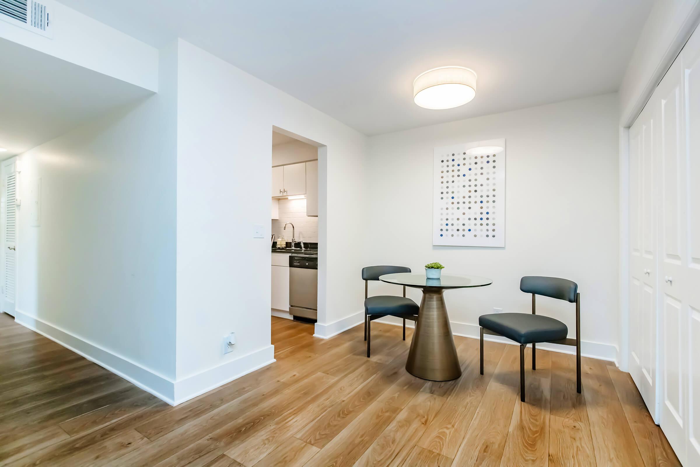 A modern, minimalist dining area featuring a round glass table with three black chairs. The space has light wood flooring and white walls. In the background, a kitchen is partially visible, and a framed artwork is mounted on the wall above the table. A contemporary light fixture hangs from the ceiling.