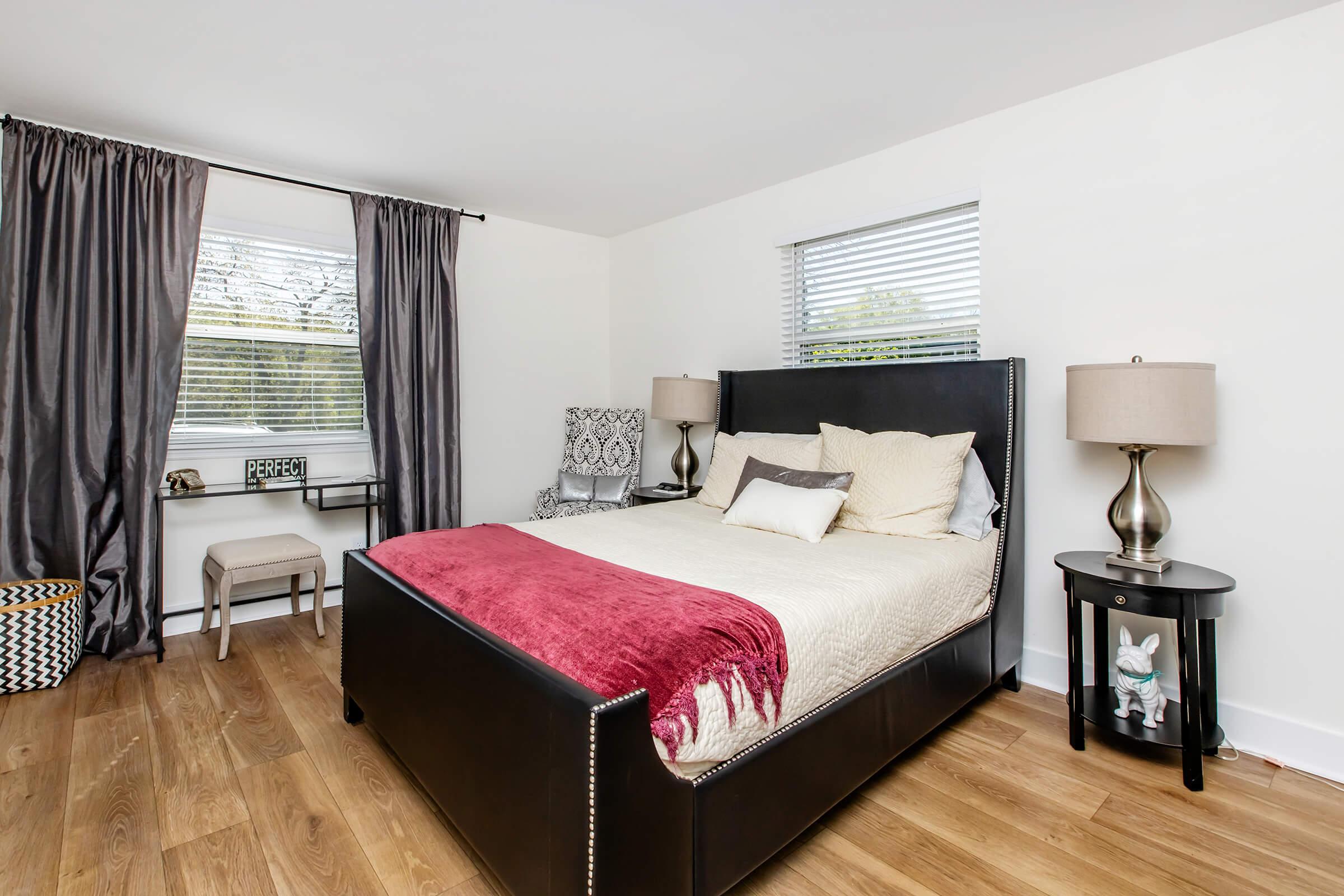 A cozy bedroom featuring a large black bed with cream and red bedding, two bedside lamps on black nightstands, light hardwood flooring, sheer white curtains, and a decorative chair in the corner. A sign that reads "Perfect" is subtly placed on a small table.