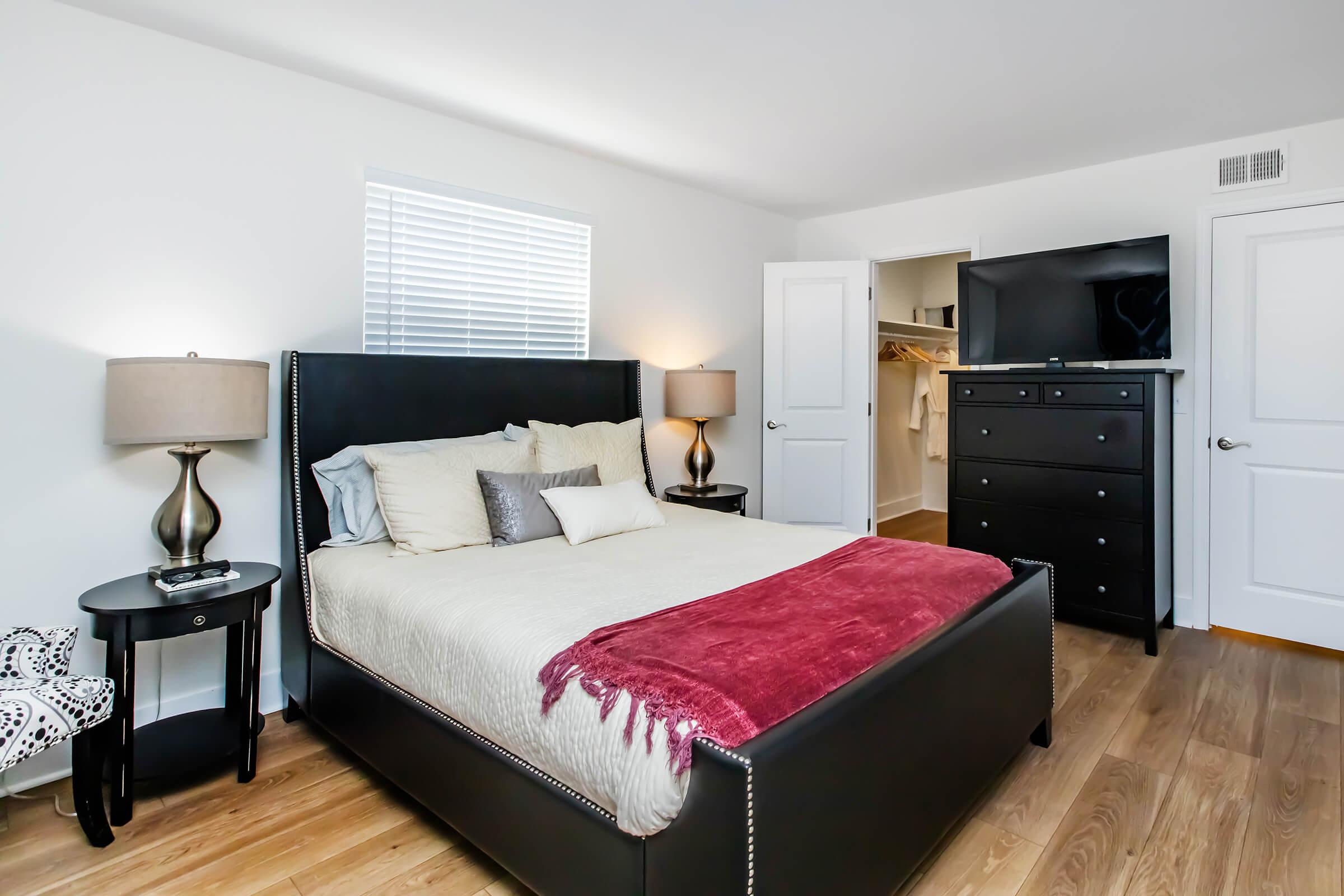 A modern bedroom featuring a black upholstered bed with a light-colored blanket, two bedside tables with lamps, a dresser, and a television. The room has wooden flooring and is illuminated by natural light from a window. A closet is visible in the background.