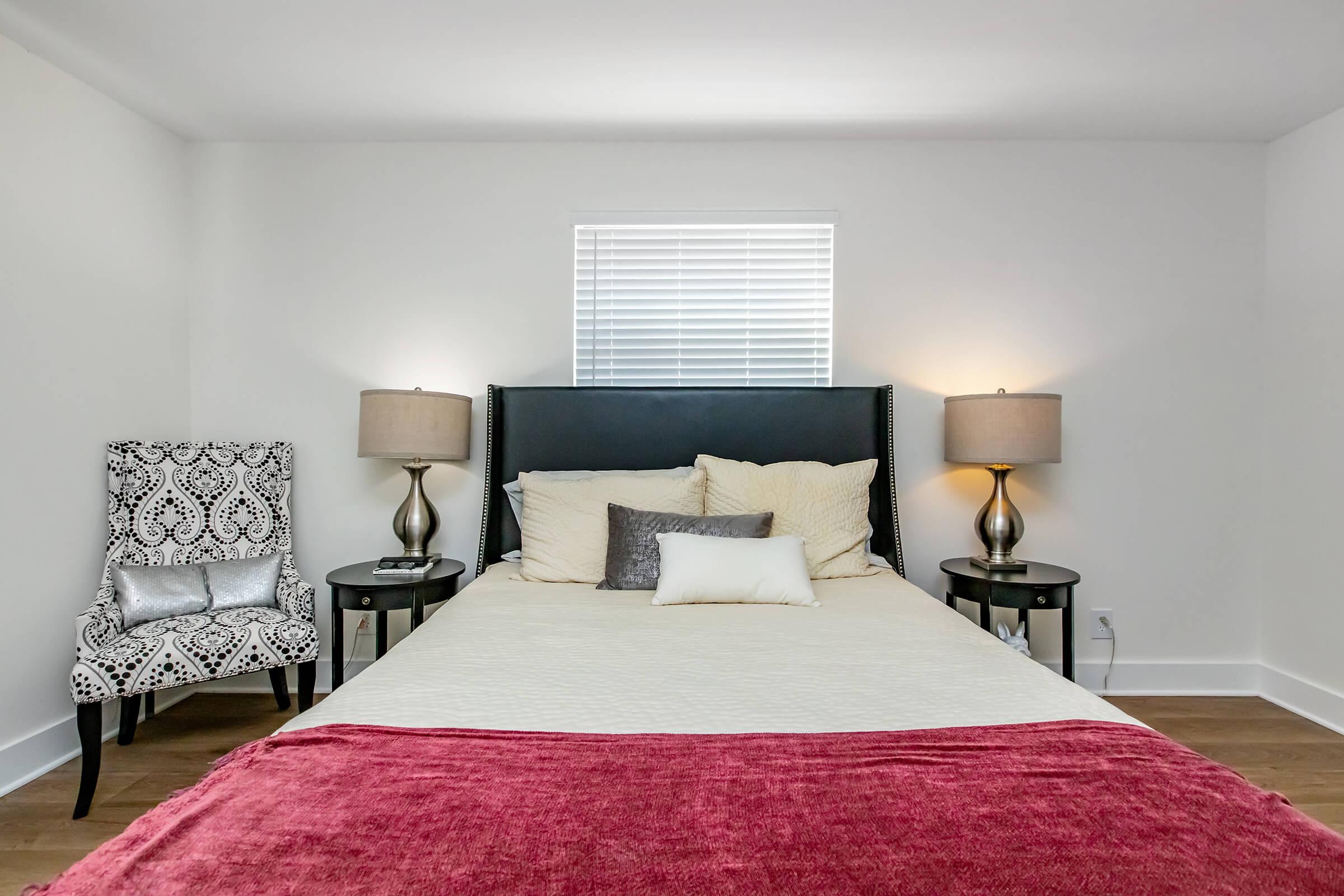 Modern bedroom featuring a black upholstered headboard with white and gray bedding. On either side of the bed, there are stylish bedside tables with lamps. A patterned armchair is positioned in the corner, and a window with blinds allows natural light into the airy, minimalist space.