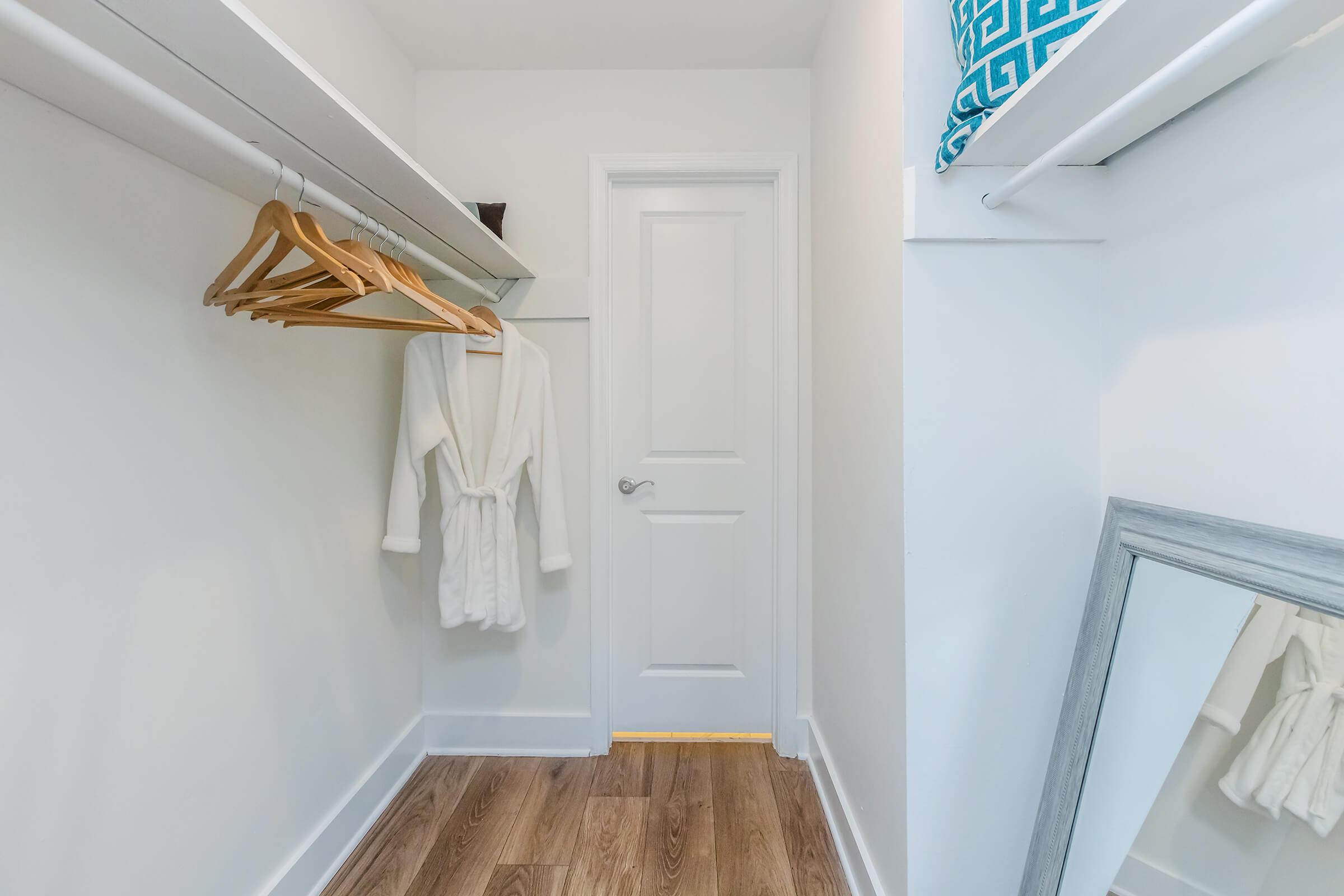A clean and organized closet featuring a white robe hanging on a wooden hanger, a mirror on the floor, and a shelf with a decorative box. The walls are painted white, and the floor is a light-colored wood, giving the space a bright and spacious feel.