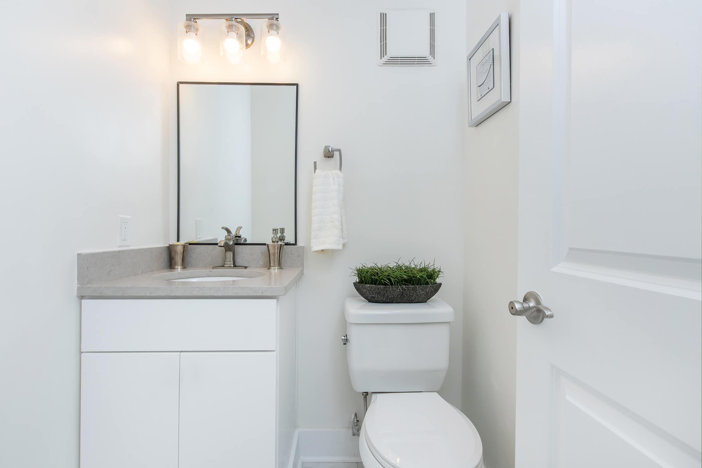 A modern bathroom featuring a sleek white vanity with a grey countertop, a rectangular mirror above it, and three pendant lights. A towel bar with a white towel is attached to the wall, and a small potted plant adds greenery. The toilet is positioned next to the vanity, against a clean white wall.
