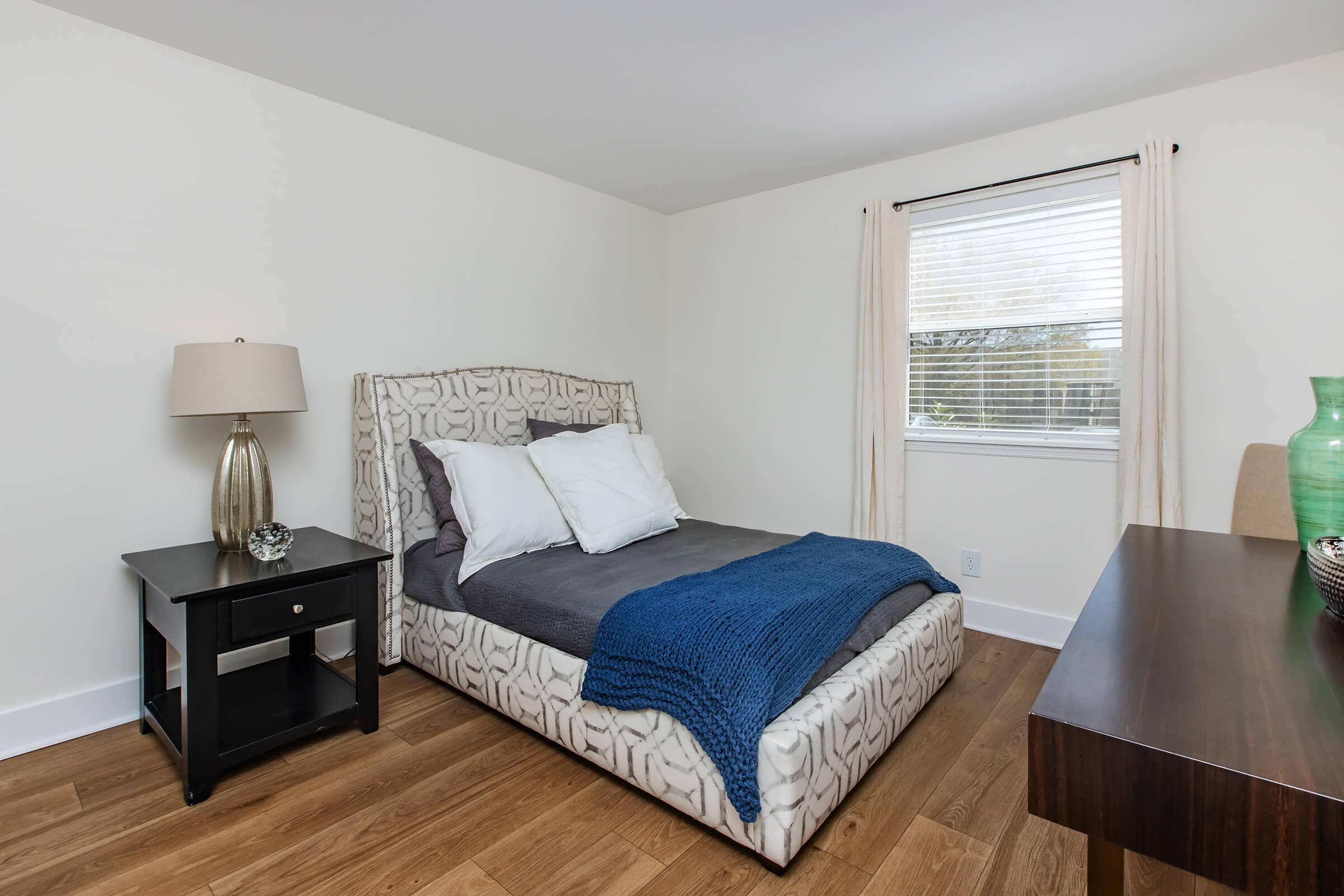 A cozy bedroom featuring a patterned bed with white pillows and a blue throw blanket. A nightstand with a lamp and decorative items is beside the bed. A wooden desk and chair are positioned against one wall, and a window with curtains allows natural light to brighten the space. The floor is made of wooden planks.
