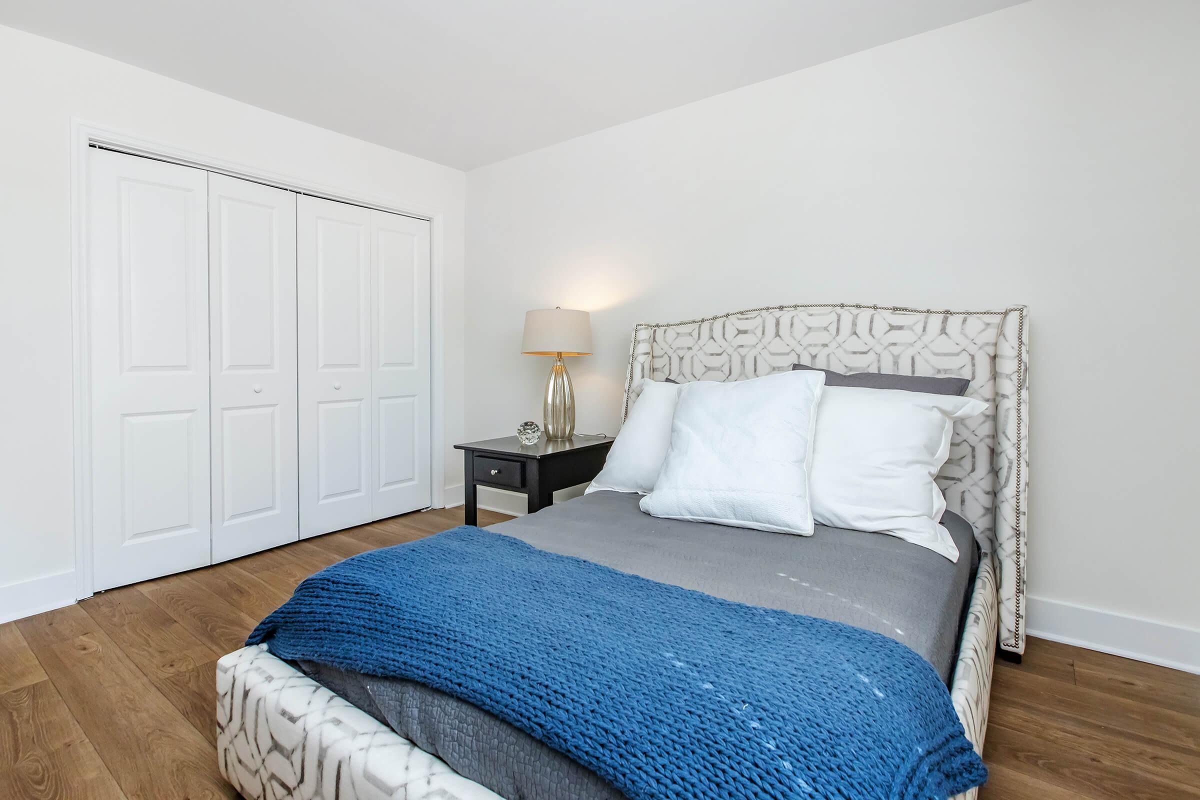 A cozy bedroom featuring a bed with a patterned headboard and gray bedding, adorned with white pillows and a blue throw blanket. A stylish nightstand is placed next to the bed, topped with a lamp. The room has light-colored walls and wooden flooring, with a closet visible in the background.