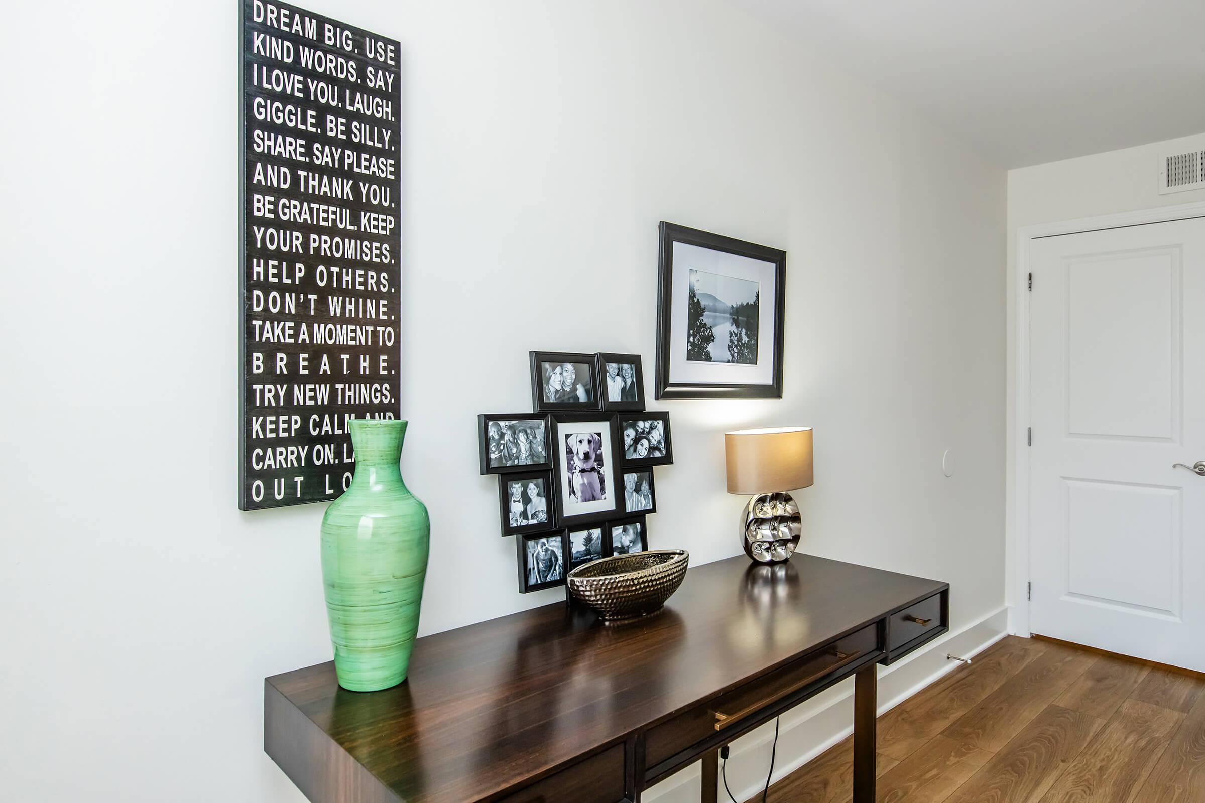 A modern entryway featuring a wooden console table with a decorative green vase, a woven bowl, and a table lamp. On the wall hangs a motivational quote sign and a framed photo collage, adding a personal touch to the space. The flooring is wooden, and the walls are painted white.