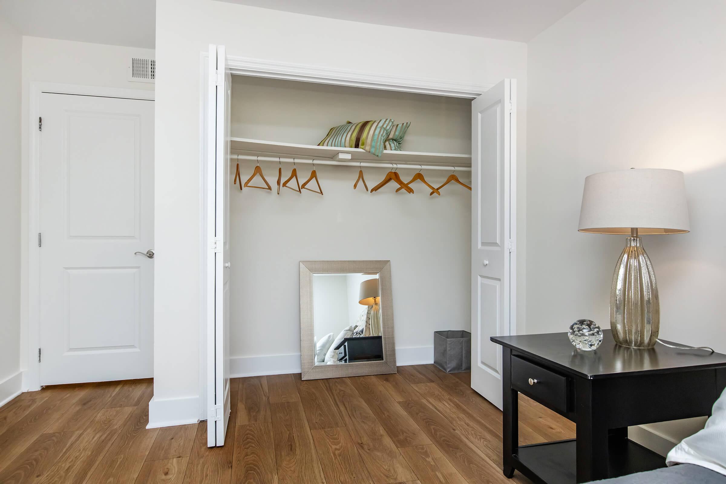 A well-lit closet with white double doors open to reveal empty wooden hangers on a shelf. Below, a mirror leans against the wall, reflecting part of the room. Next to the closet, a small black nightstand holds a decorative glass orb and a stylish lamp, with warm wooden flooring completing the contemporary look.