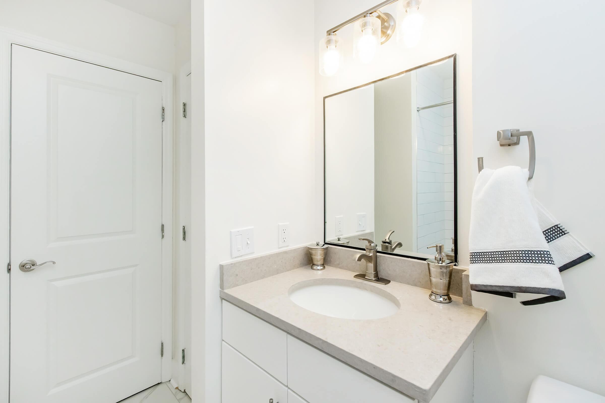 A clean and modern bathroom featuring a white vanity with a round sink and brushed nickel faucet. Above the sink, there is a rectangular mirror with three light fixtures. A white towel with a black border hangs from a chrome holder, and a closed door is visible in the background. The walls are painted white, creating a bright atmosphere.