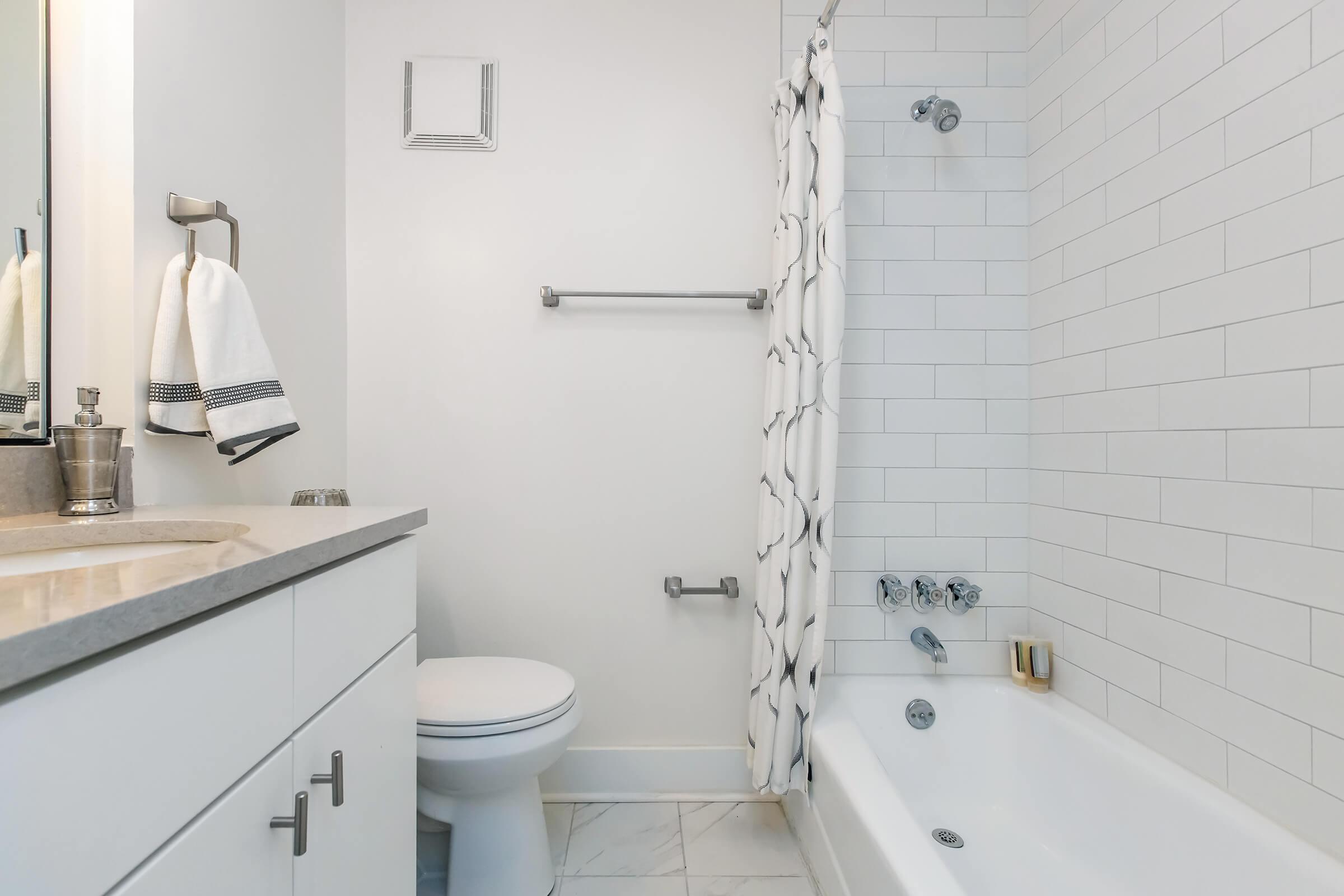 A modern bathroom featuring a white tiled shower area with a showerhead, a bathtub, a toilet, and a sink with a light-colored countertop. Towels are hanging on a towel rack, and the walls are painted in soft neutral tones. A minimalist design emphasizes a clean and spacious look.