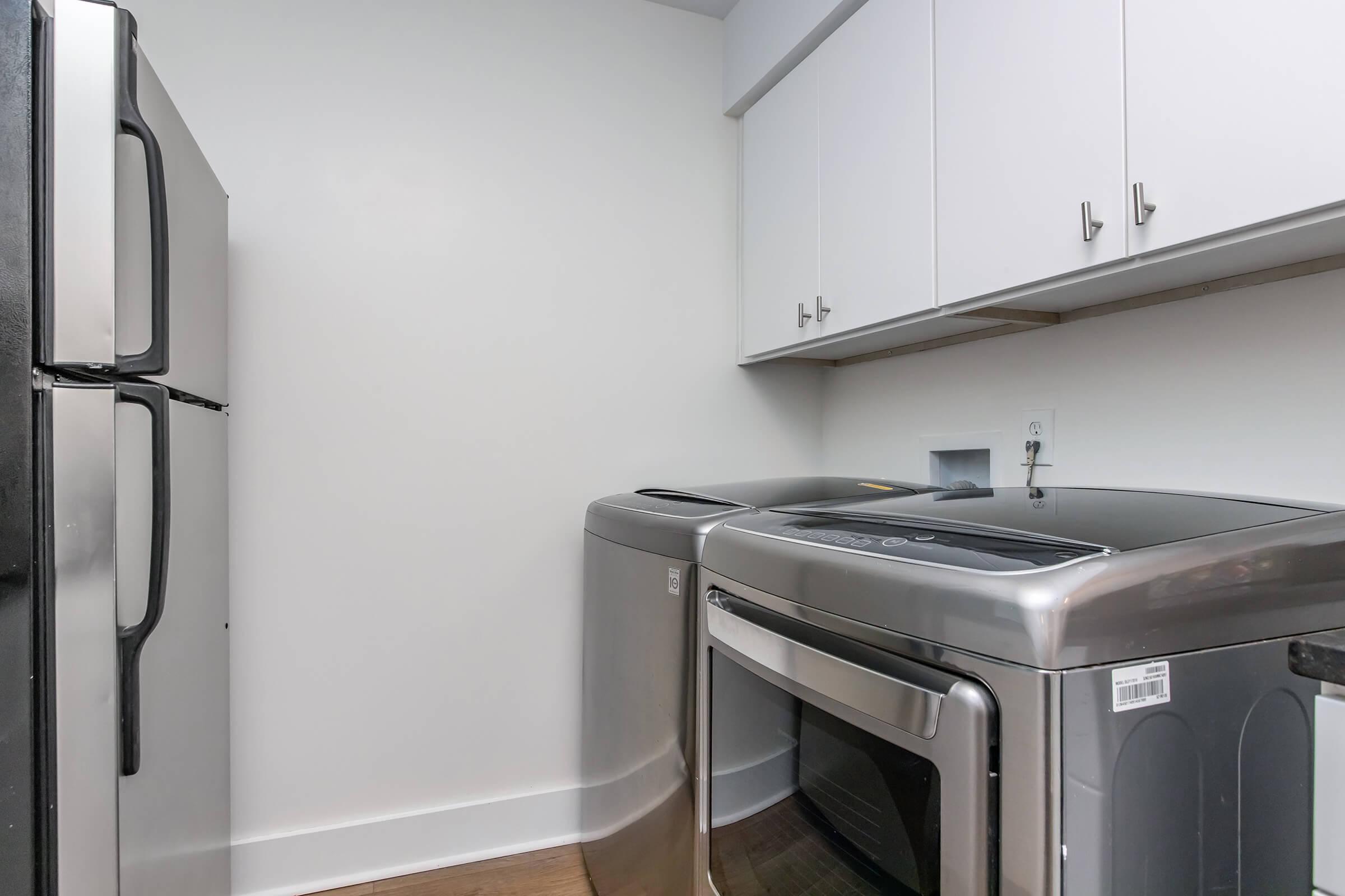 A laundry room featuring a silver washing machine and dryer side by side, with a black refrigerator in the corner. Above the appliances, there are white cabinets for storage. The walls are painted white, and the floor is made of light wood. The space is well-lit and organized.