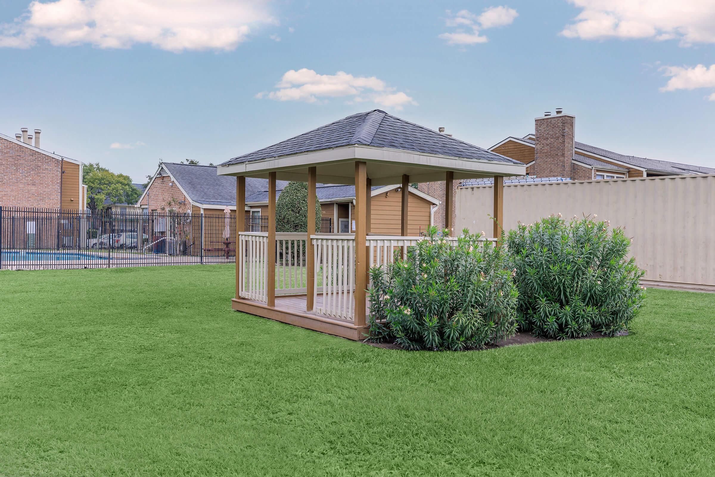 A wooden gazebo with a sloped roof sits in a green grassy area, surrounded by shrubs. In the background, there are residential buildings and a fenced swimming pool area under a clear blue sky.