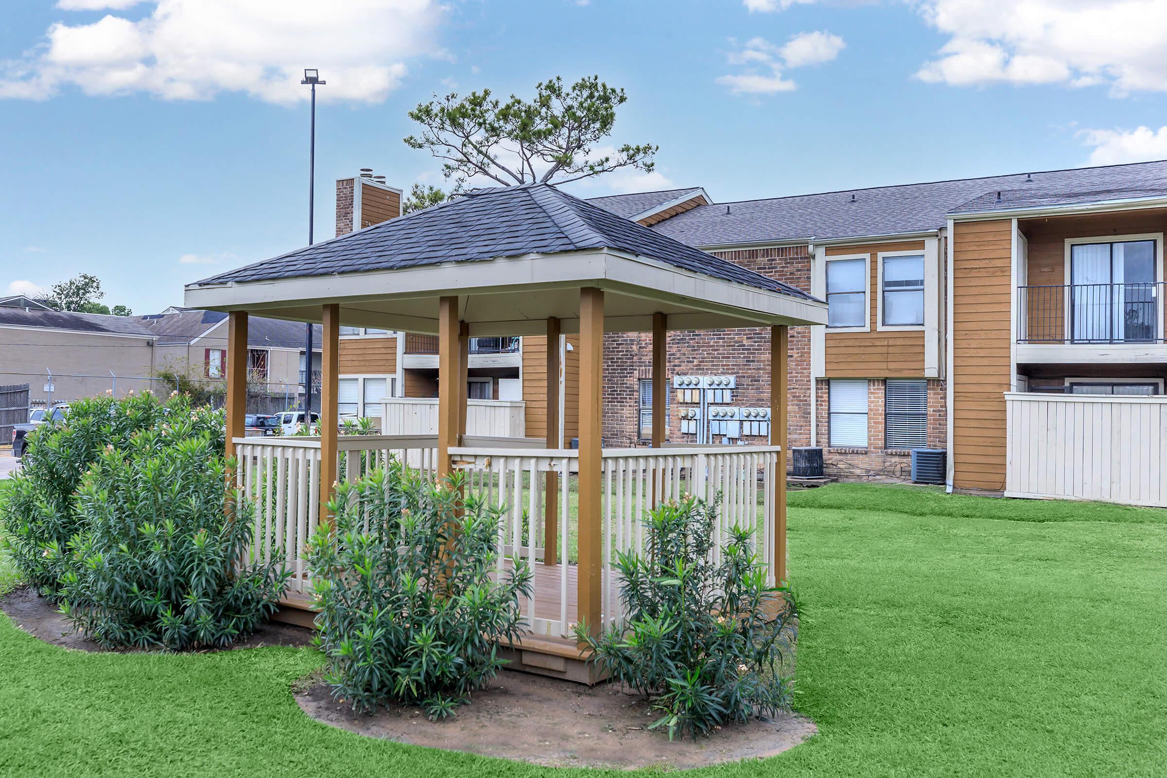 A wooden gazebo with a dark shingle roof situated in a landscaped area featuring lush green grass and shrubs. In the background, there are residential buildings with balconies, under a clear blue sky with a few clouds. The scene is serene and inviting, perfect for relaxation.