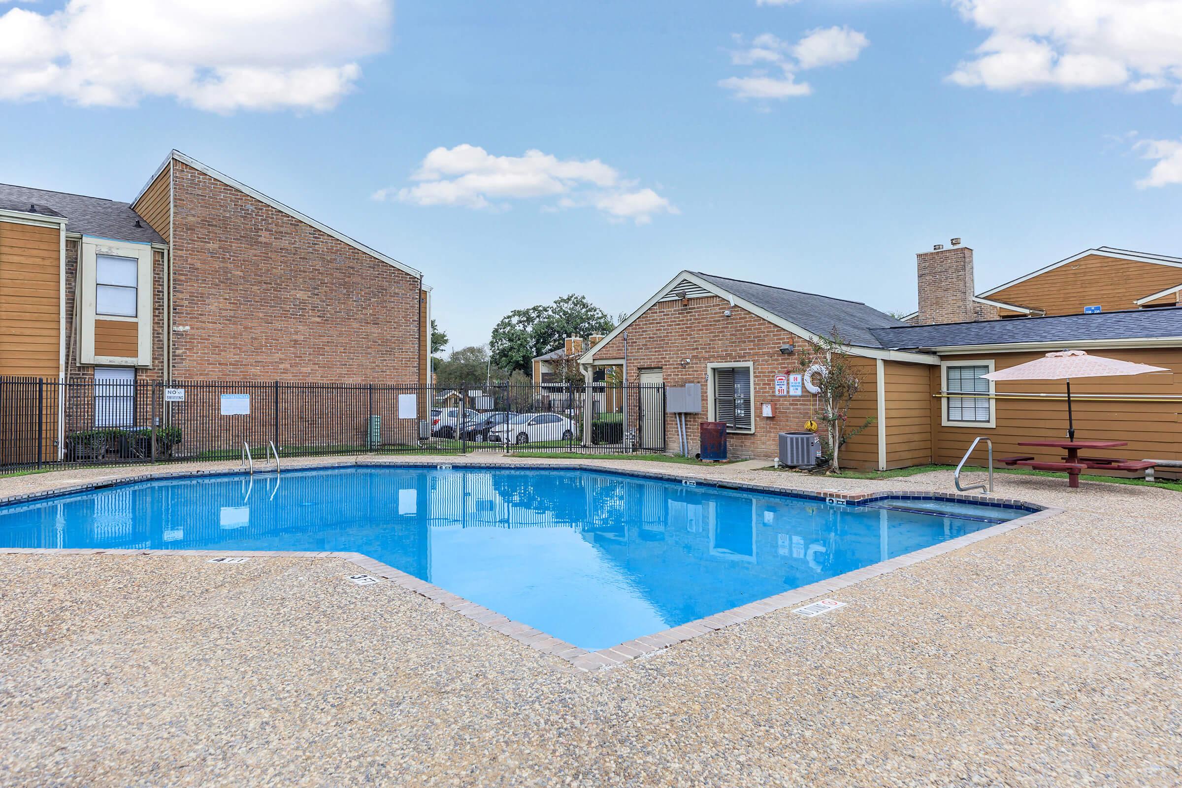 A clear blue swimming pool surrounded by a stone deck, with a picnic table and an umbrella nearby. Various buildings with orange and brick exteriors are in the background, under a blue sky with a few clouds. A fence encloses the pool area.