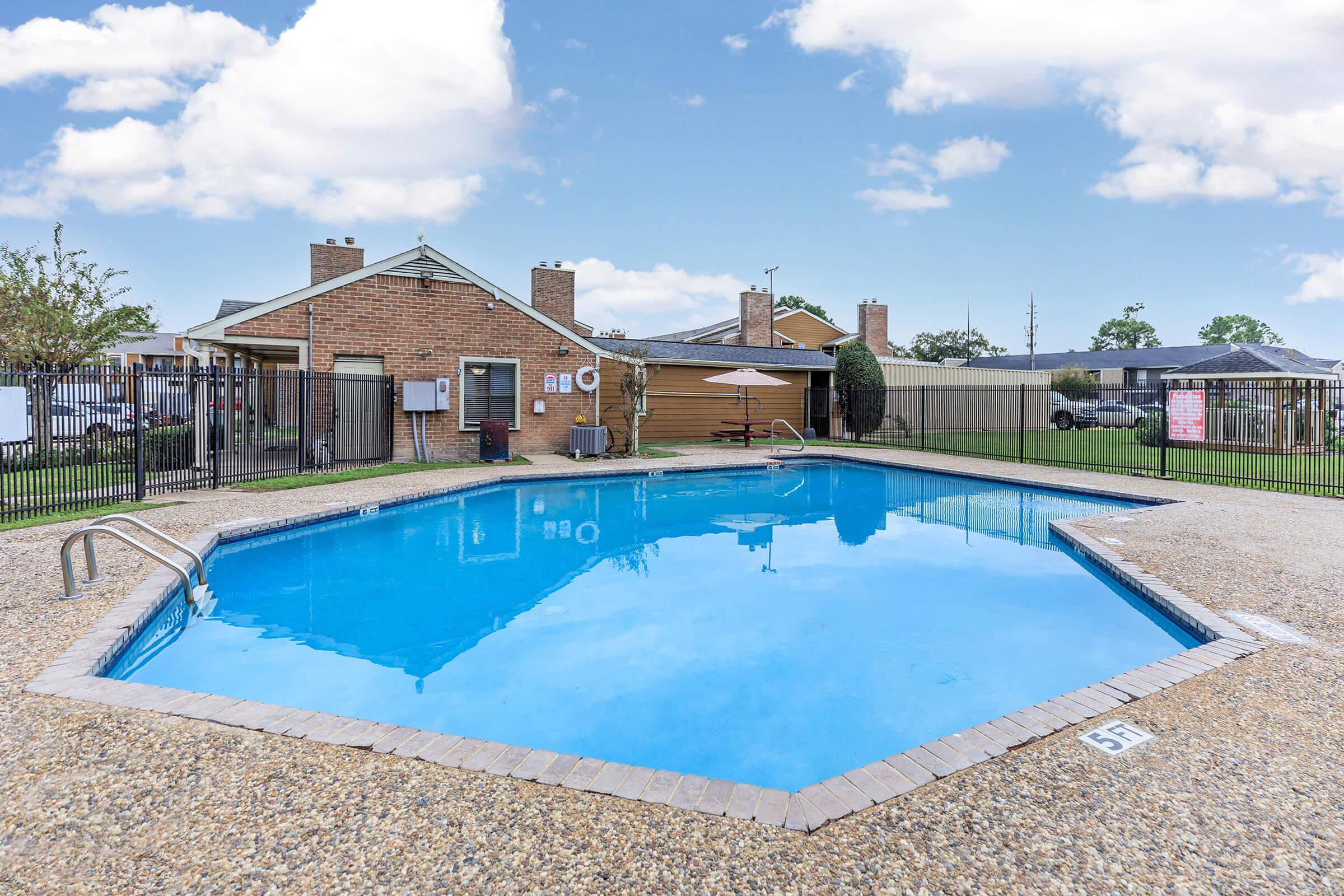 A rectangular swimming pool with clear blue water surrounded by a textured concrete deck. In the background, there are brick buildings with a fence and trees. A single umbrella is set up near the pool, and the sky above is partly cloudy.