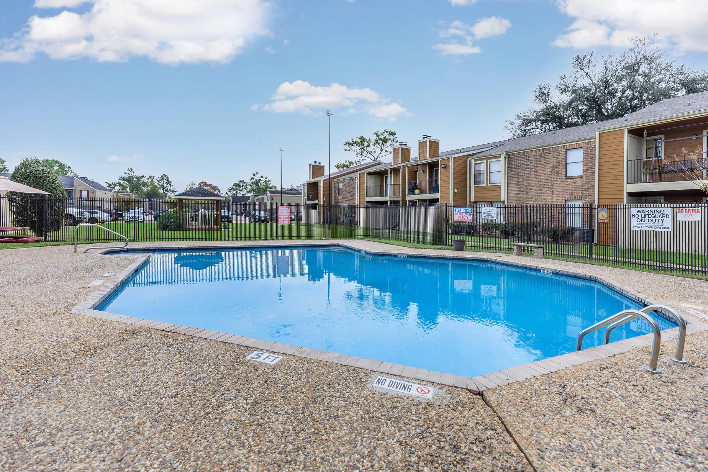 A clear blue swimming pool surrounded by a concrete deck and grassy area, with two apartment buildings in the background. A diving board is visible at one end, and pool safety markings are present on the deck. The scene is bright and inviting, showcasing a recreational outdoor space.