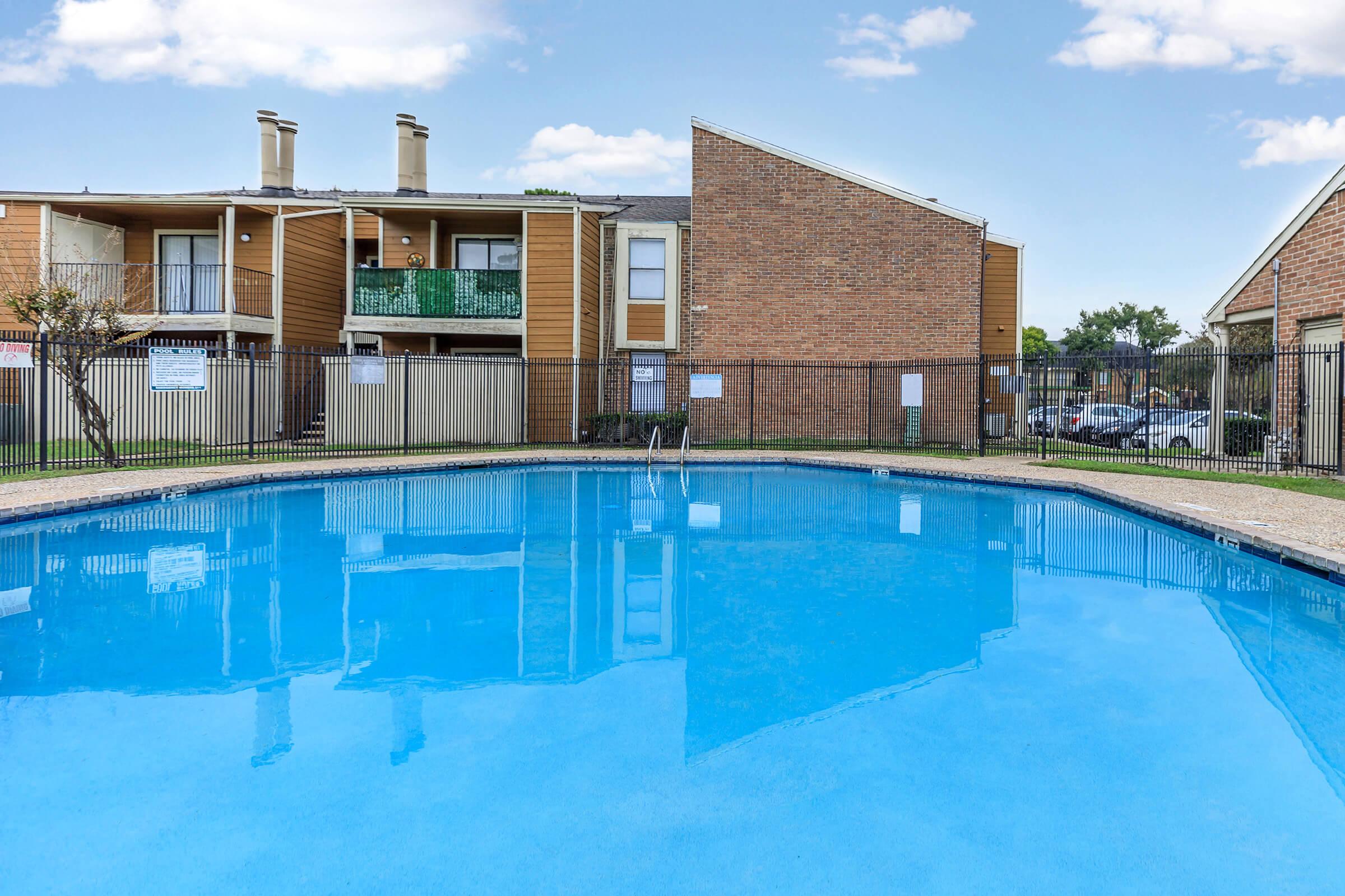 A clean and inviting swimming pool surrounded by a fenced area. In the background, there are two two-story apartment buildings featuring brick and wood siding, with green balconies and parking visible. The sky is clear with a few clouds, creating a bright atmosphere.
