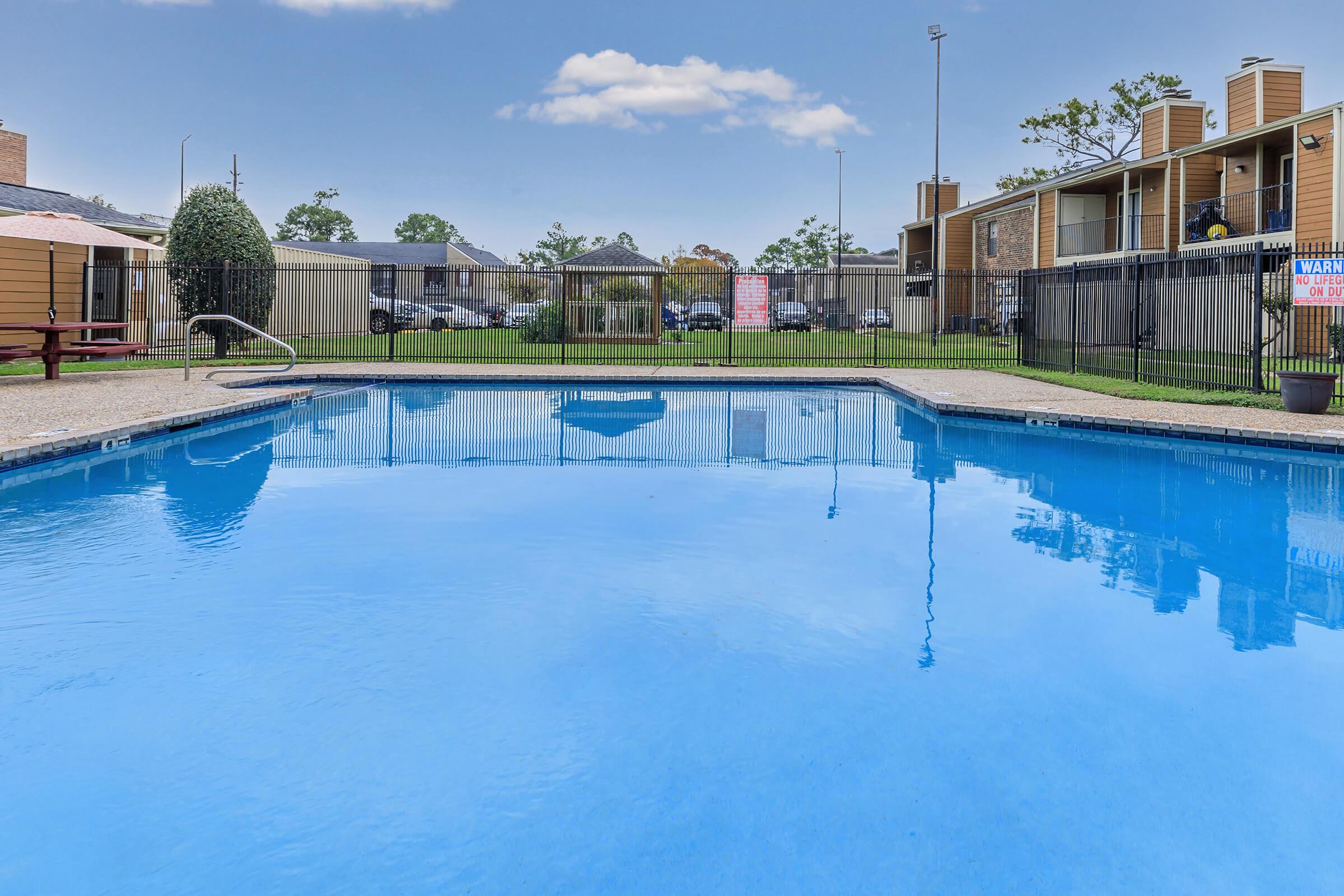 A clear blue swimming pool surrounded by a fence, with lounge chairs nearby. In the background, there are several buildings and a grassy area. A sign can be seen, along with trees and a cloudy sky above.