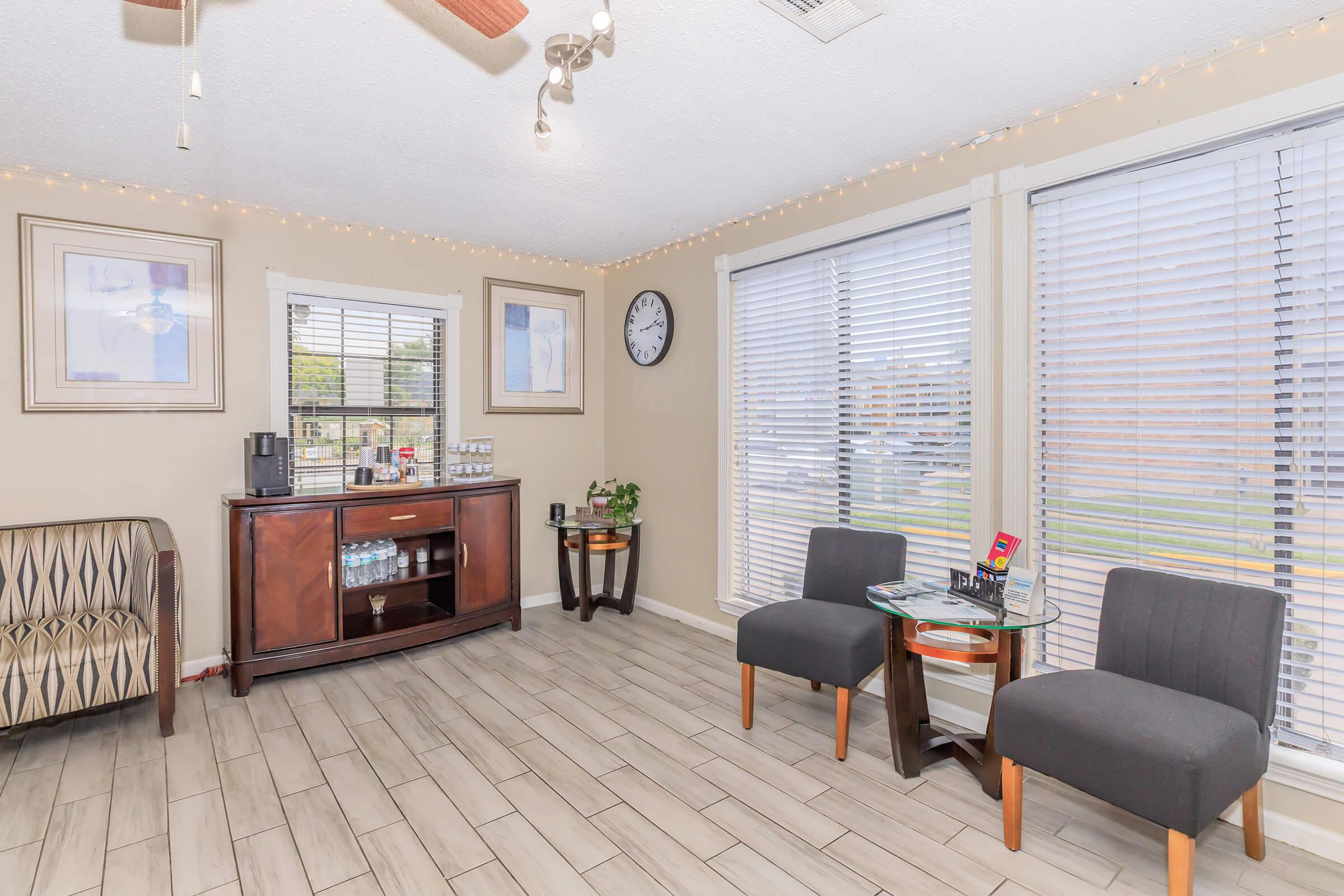 A cozy sitting area featuring two gray chairs and a small glass table, with a wooden cabinet displaying beverages and snacks. Sunlight streams through large windows adorned with blinds. There’s a wall clock and decorative frames enhance the warm atmosphere, suggesting a welcoming space for relaxation.