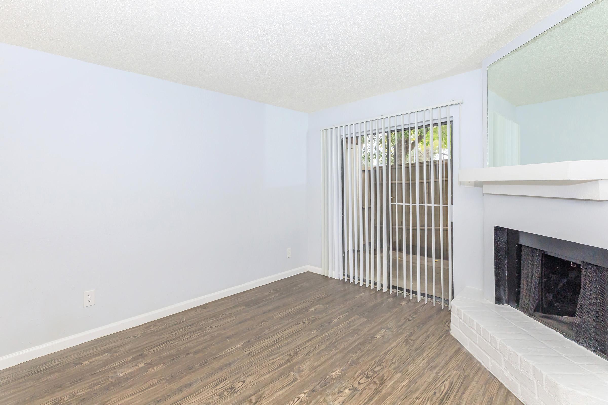 Interior view of a room featuring light blue walls, a white brick fireplace, and a set of vertical blinds covering a sliding glass door. The floor has a wood-like finish, and the space appears empty and well-lit.