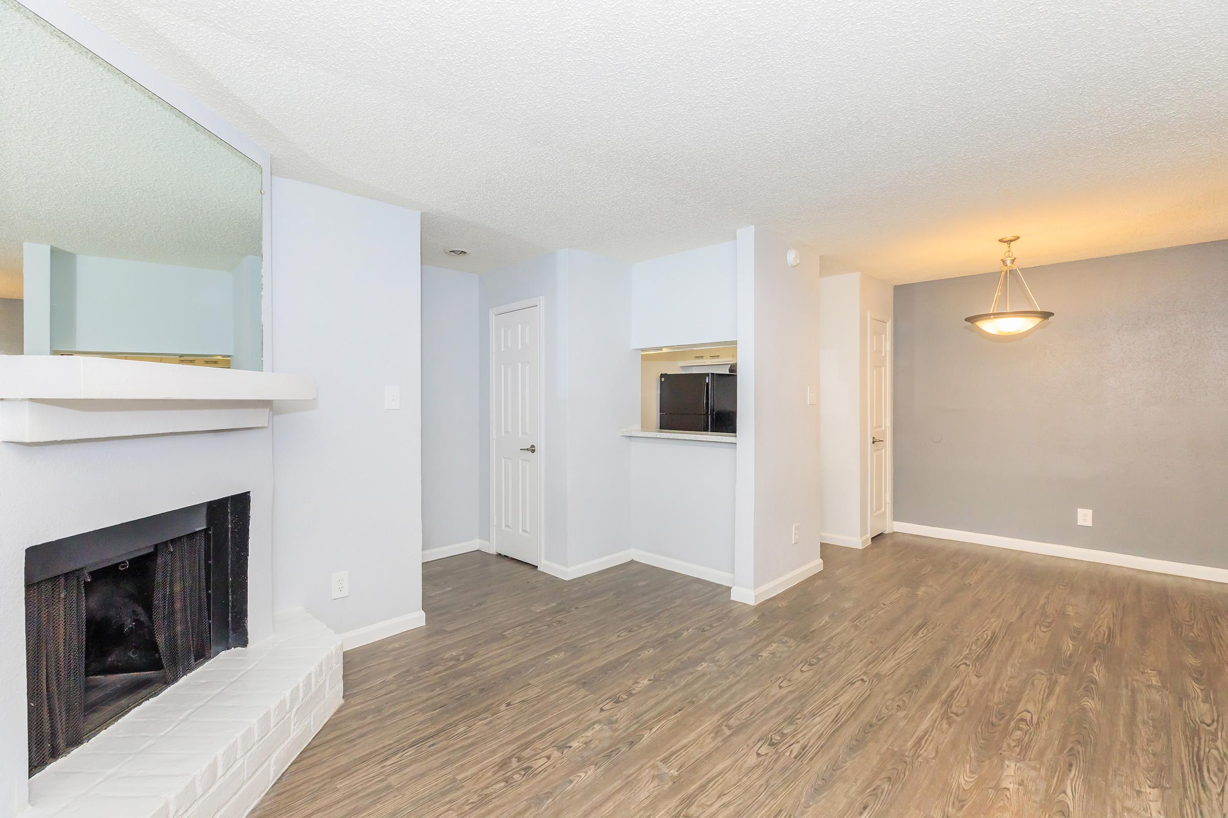 Open living space featuring a fireplace, light gray walls, and wood-style flooring. To the right, there's a small kitchenette with an open pass-through. A ceiling light fixture hangs above the dining area, adding warmth to the room. The overall design is modern and inviting.