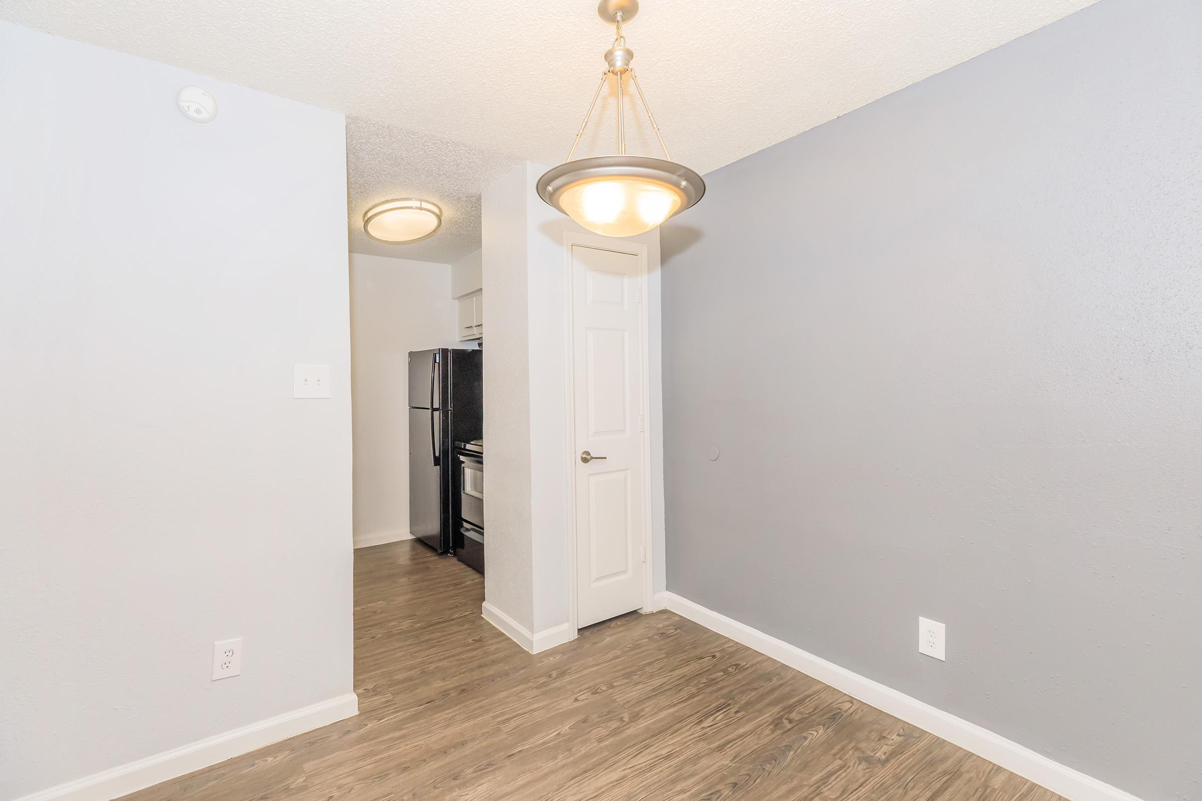Interior view of a small, modern kitchen area featuring gray walls, a hanging light fixture, and laminate flooring. The space includes a doorway leading to an adjacent area, with a black refrigerator visible in the background. The decor is minimal and clean, reflecting a contemporary design.