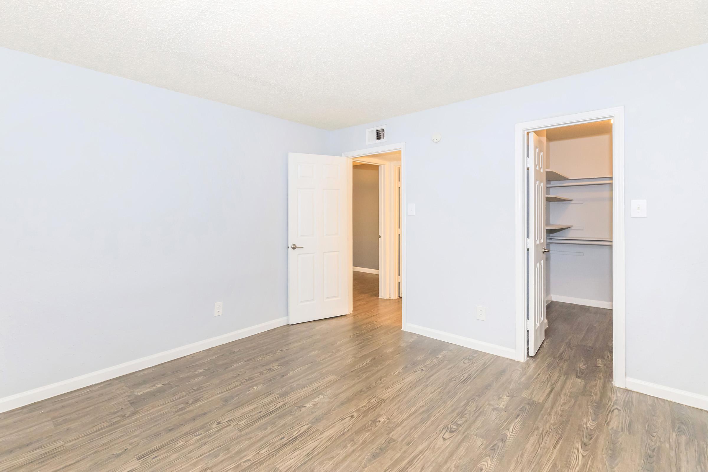 Empty room with light blue walls and a wooden floor. One door leads to another room, and a closet with shelves is visible on the right. The room appears bright and uncluttered, featuring a ceiling with a textured finish.