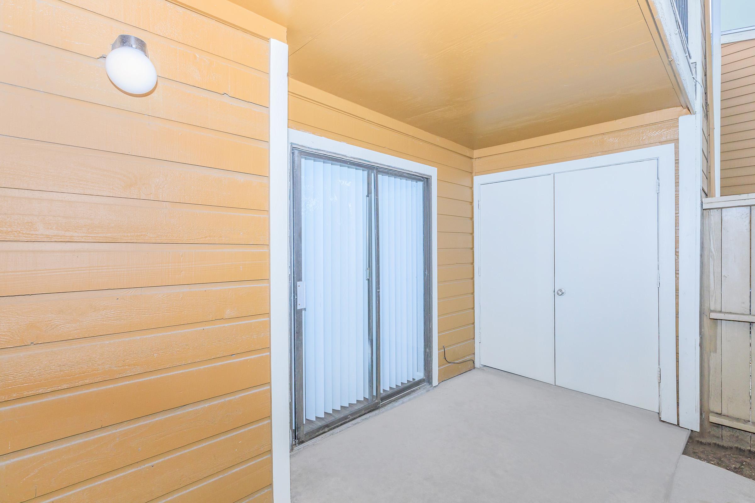 A covered outdoor space with beige wooden siding, featuring a sliding glass door and a white door at the end. The ceiling is painted orange, and there's a light fixture on the wall. The floor is concrete, creating a clean and open area.