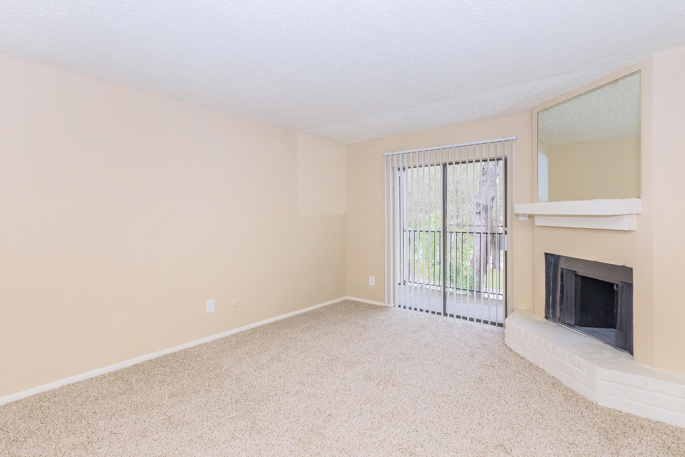 A spacious, empty living room featuring neutral beige walls and carpet. There is a large window with vertical blinds allowing natural light, and a fireplace on one wall. The room has a cozy, inviting atmosphere, suitable for various furniture arrangements.