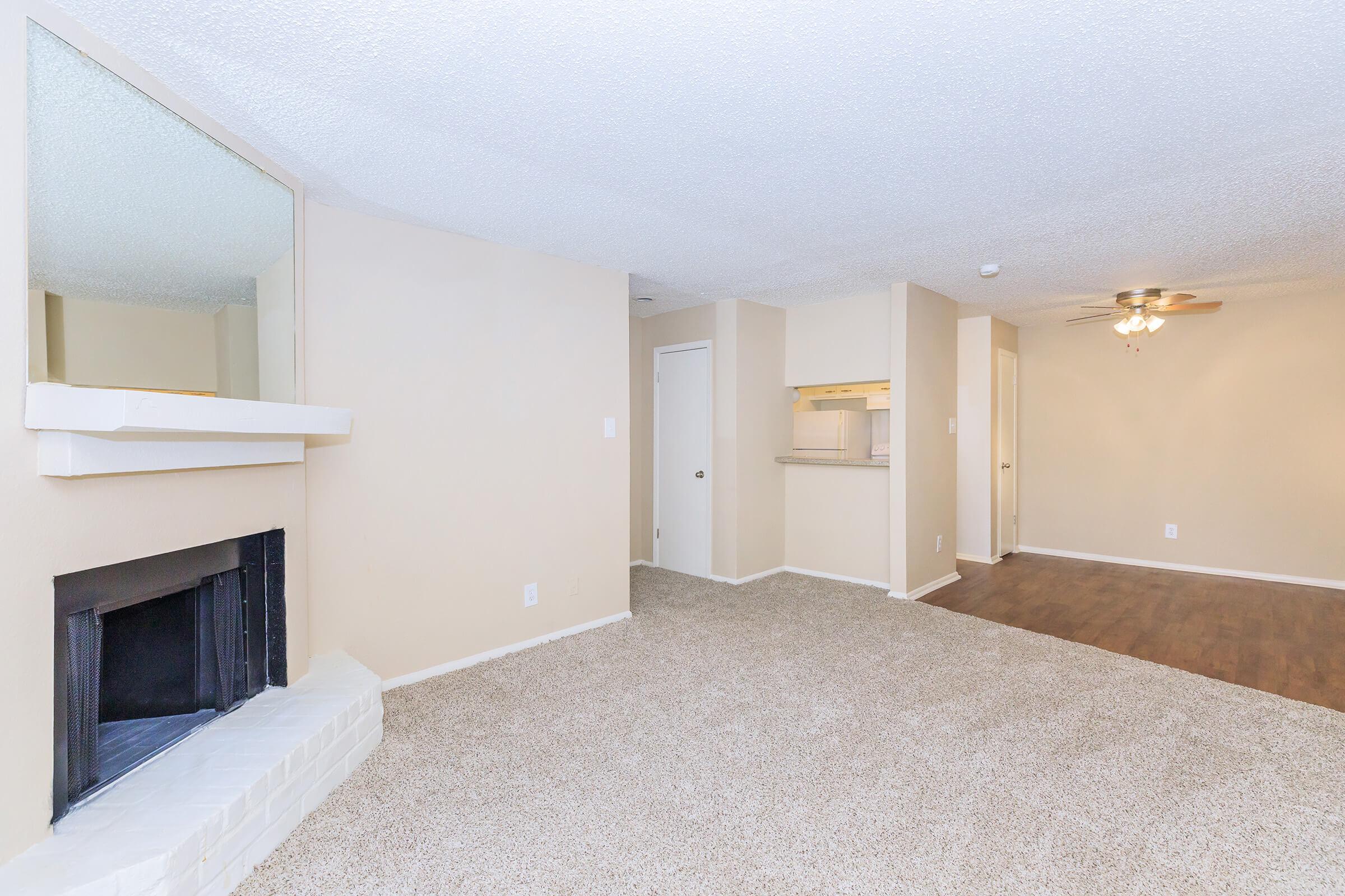 A spacious living area featuring light-colored walls and carpet, a cozy white brick fireplace, and an open plan leading into a kitchen. There is a door on one side, and a ceiling fan provides airflow. The space is well-lit and ready for furnishings, creating a warm and inviting atmosphere.